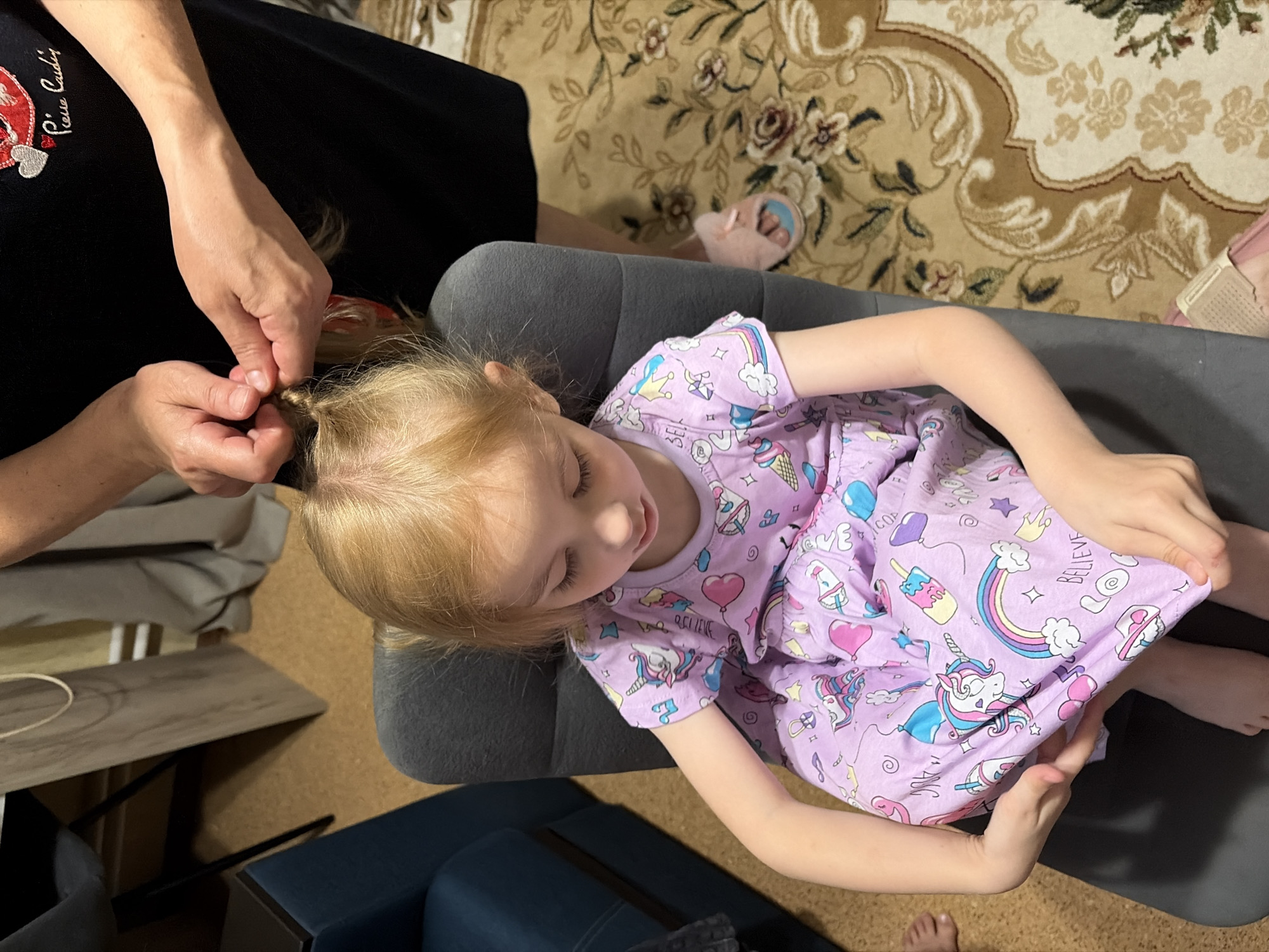 A young girl with blonde hair, wearing a purple dress with unicorn and ice cream motifs, is sitting on a grey chair. Her eyes are closed as an adult, whose hands are visible, braids her hair. The background shows a textured carpet with floral patterns, a dark blue armchair, and a wooden surface. The scene appears to be indoors, possibly a home, with no clear indication of its specific location within Tambov, Russia.