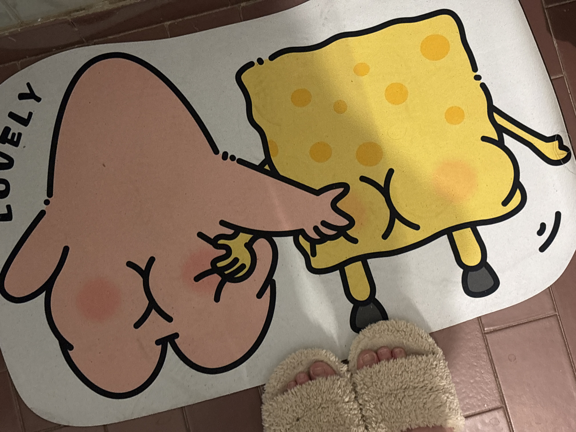 A cartoon mat depicts Patrick Star touching SpongeBob SquarePants' bottom. The mat sits on a tiled floor. A pair of fluffy slippers with visible toes rest on the mat. The location is not determinable from the image.