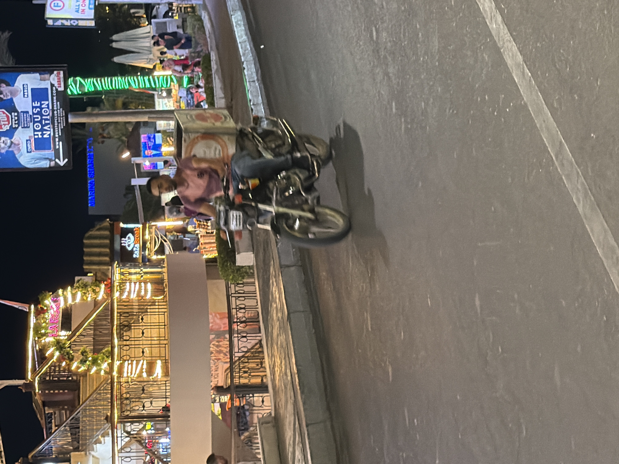 A man is riding a motorcycle down a street at night in Sharm El Sheikh, Egypt. The man is wearing a pink t-shirt and blue jeans. The motorcycle has a large box on the back, possibly for deliveries. In the background, there are several buildings and signs, including one for "HOUSE NATION" and another for "MARINA SHARM." The street is wet and reflective, suggesting it has recently rained or been washed. Lights from the buildings and signs illuminate the scene.