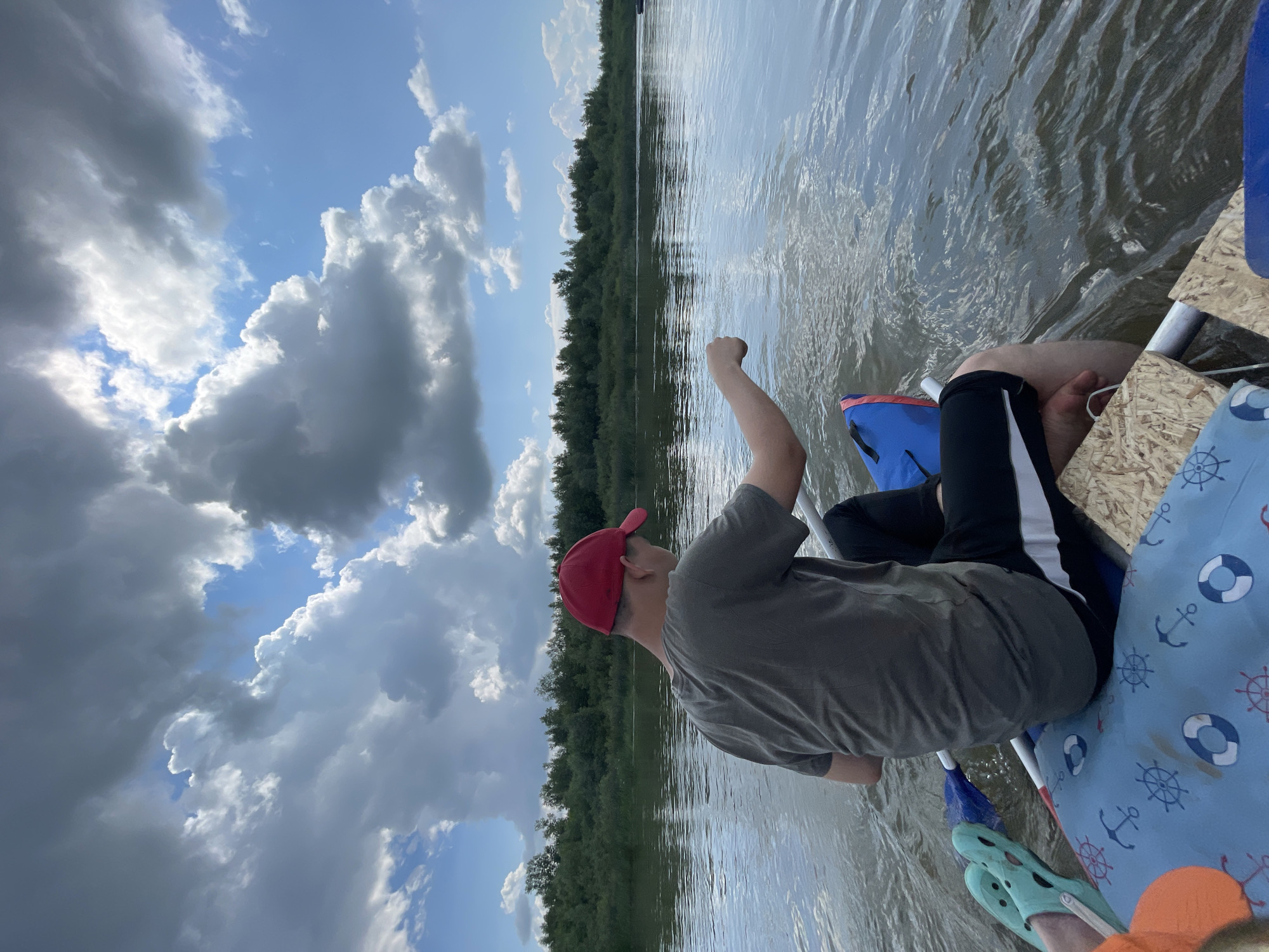 A person wearing a red cap and a grey t-shirt is seated in a raft on a body of water. The person is facing away from the camera, and their right arm is extended, with their hand appearing to be in the water. The raft has a blue and red bag and a blue mat with anchor and life preserver patterns. Light blue Crocs are visible on the raft. The water is calm with ripples. In the background, a line of dense green trees runs along the horizon. The sky is a bright blue with scattered, dramatic clouds, some dark and some white and fluffy. The scene suggests an outdoor recreational activity on a river or lake under a partly cloudy sky. The image appears to have been taken from the perspective of someone also in the raft. The general location is described as the city of Нежинка, Russia.