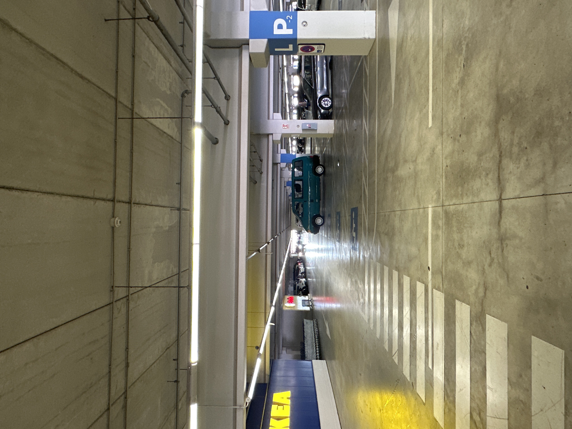 The image depicts the interior of a multi-level parking garage at IKEA in Alfafar, Spain. The concrete ceiling, supported by columns, features exposed conduits and fluorescent lighting. The floor is marked with white lines indicating parking spaces and a pedestrian crossing. In the foreground, a pedestrian crossing is visible with white stripes. To the left, a section of the IKEA store's blue facade with yellow lettering is present. In the middle ground, a teal van is parked. Further back, several cars are parked in designated spots, and shopping carts are lined up near the store entrance. A blue and white sign with "L P -2" is visible on a column, indicating parking level. The environment is illuminated by artificial lighting, creating a utilitarian atmosphere. There are no people visible in the immediate scene.