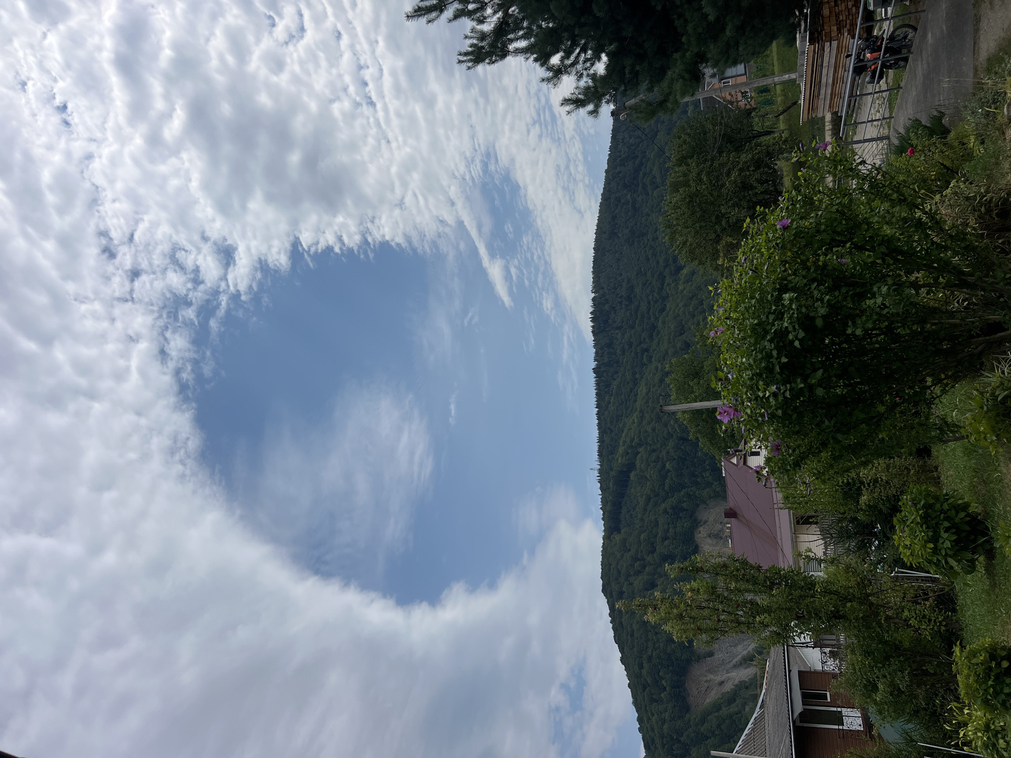 In Yaremche, Ukraine, a picturesque scene unfolds under a sky dominated by dramatic clouds. A large, circular gap in the clouds reveals a bright blue sky, creating a striking contrast with the surrounding white formations. Below, a lush, green hillside is densely covered with trees, forming a verdant backdrop. A prominent landslide scar is visible on the left side of the hill, a stark reminder of natural forces. In the foreground, several houses with reddish-brown roofs are nestled amidst abundant greenery. A vibrant bush with purple flowers adds a splash of color to the scene. To the right, a motorcycle is parked on a concrete path, adjacent to a stack of lumber. The overall impression is one of serene natural beauty tinged with the evidence of geological activity.