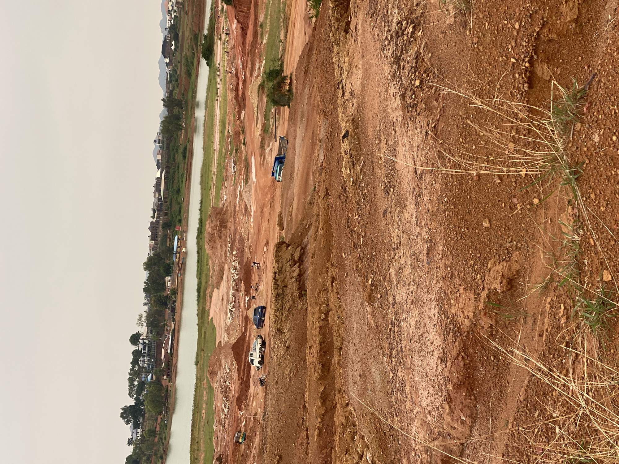 The image captures a landscape in Jos, Nigeria, characterized by a large body of water in the background, bordered by a developed area with buildings and vegetation. In the foreground, the terrain is predominantly red, rocky earth, indicating a mining or construction site. Several vehicles, including a blue truck, a white pickup truck, a blue car, and a motorcycle, are present in the middle ground, suggesting ongoing activity. A few individuals are visible in the distance, engaged in what appears to be work. The overall impression is a scene of development and resource extraction in a semi-urban environment.