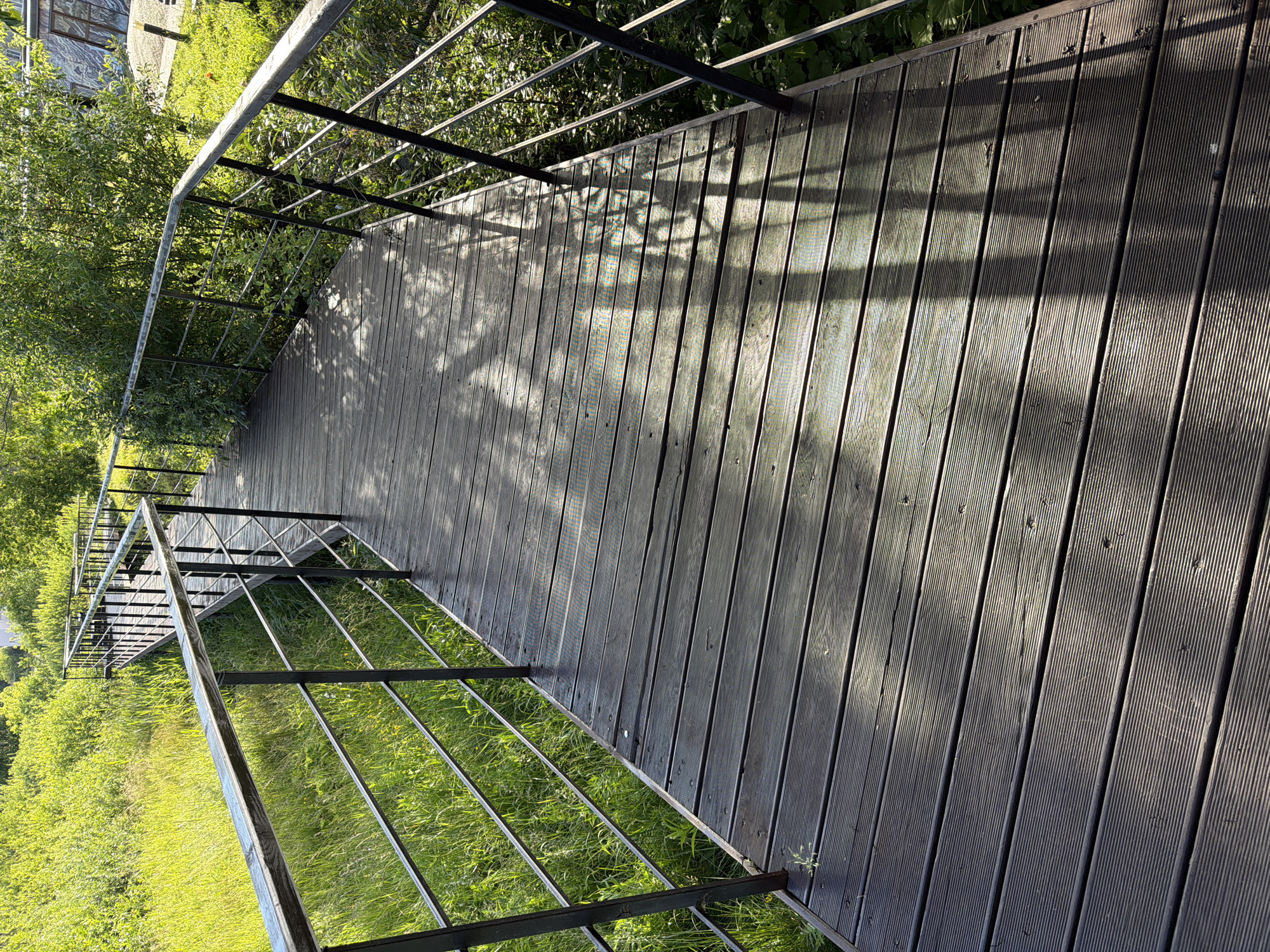 A wooden boardwalk with metal railings curves through a grassy area with trees. The boardwalk is constructed of dark, weathered wooden planks. The railings are made of black metal with a silver handrail. Sunlight filters through the leaves of the trees, casting dappled shadows on the boardwalk. The surrounding environment is lush and green, with tall grass and dense foliage. The scene suggests a park or a natural area within a city.