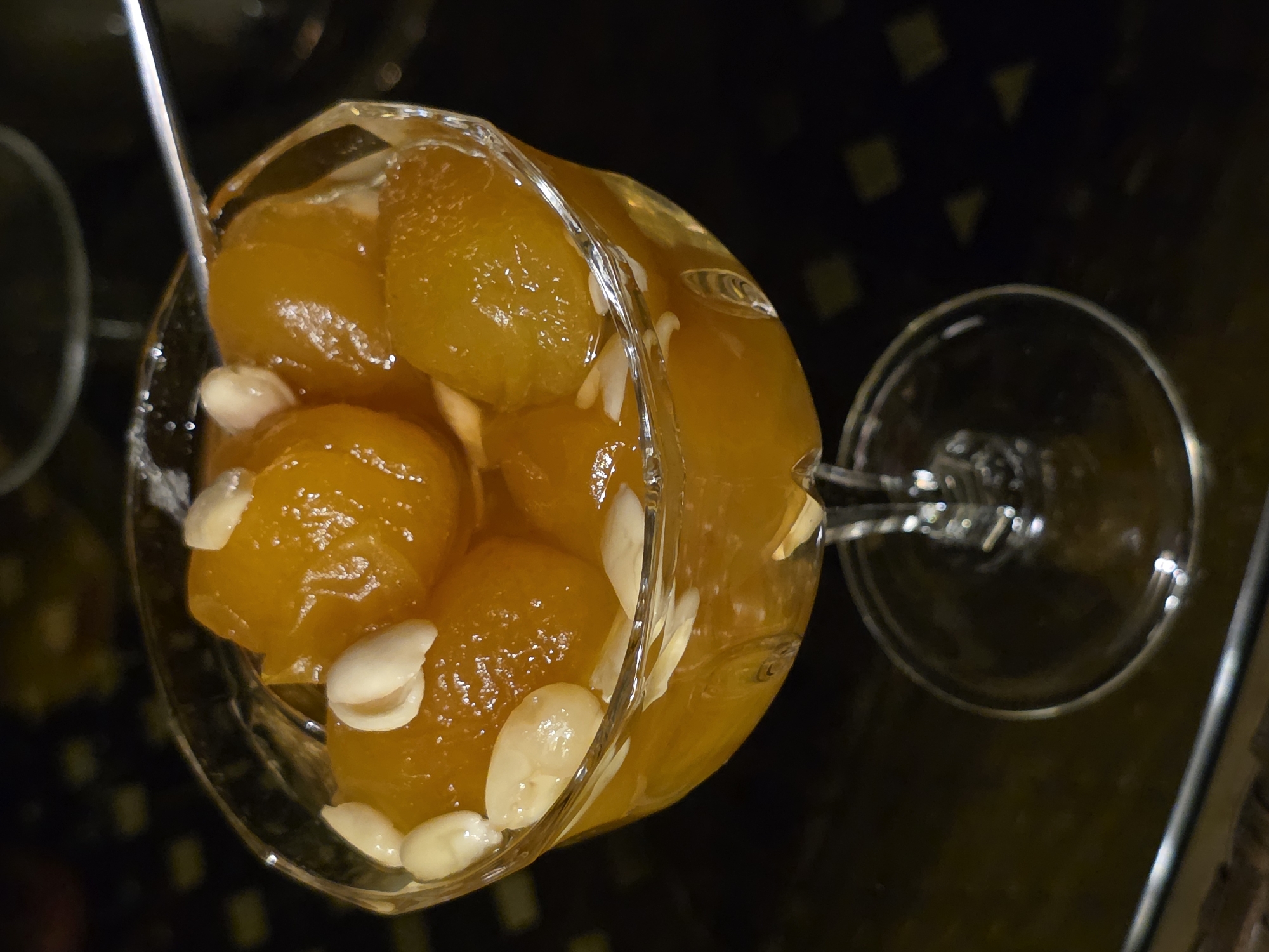 In a dimly lit environment, a clear glass coupe is filled with golden-hued candied fruits, likely apricots or similar, studded with slivered almonds. The dessert is positioned on a dark, patterned surface, possibly a tablecloth or tray, which creates a contrast with the bright contents of the glass. A silver spoon is partially visible, resting on the rim and dipping into the candied fruits. The overall impression is one of a sweet delicacy presented for consumption. There is no information in the image to suggest a location in Buzovna, Azerbaijan.