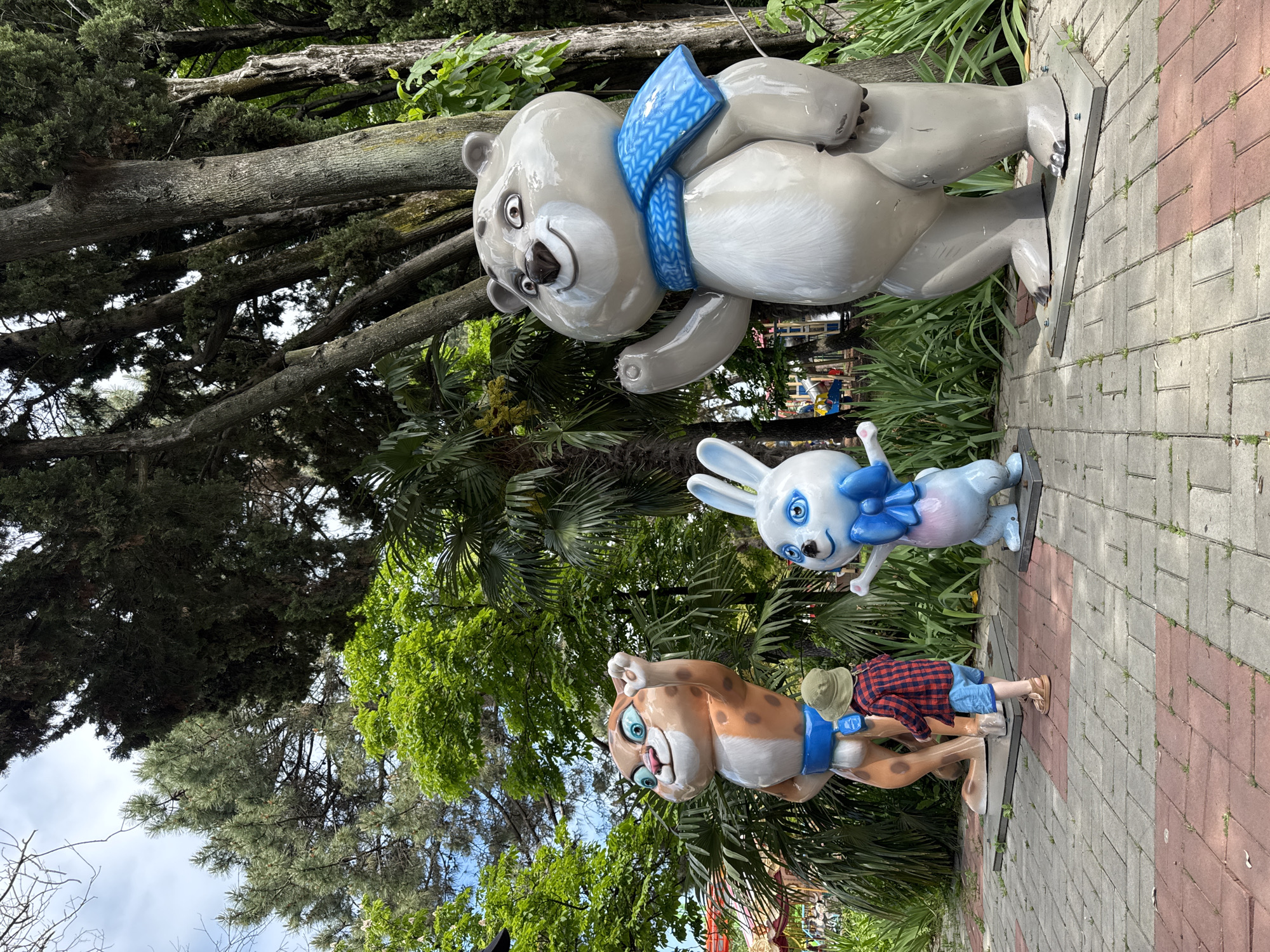 The image shows three cartoonish animal statues and a child in a park or amusement area, likely in Sochi, Russia. The statues are arranged in a row: a bear, a rabbit, and a leopard, each wearing a blue scarf or bow. The statues appear to be made of a shiny material, possibly plastic or resin. The bear is light gray, the rabbit is white, and the leopard is tan with orange spots.
A young child wearing a red and black plaid shirt, blue shorts, and a green hat is partially obscured by the leopard statue. The child is standing on a brick-paved walkway. Lush green trees and vegetation fill the background.