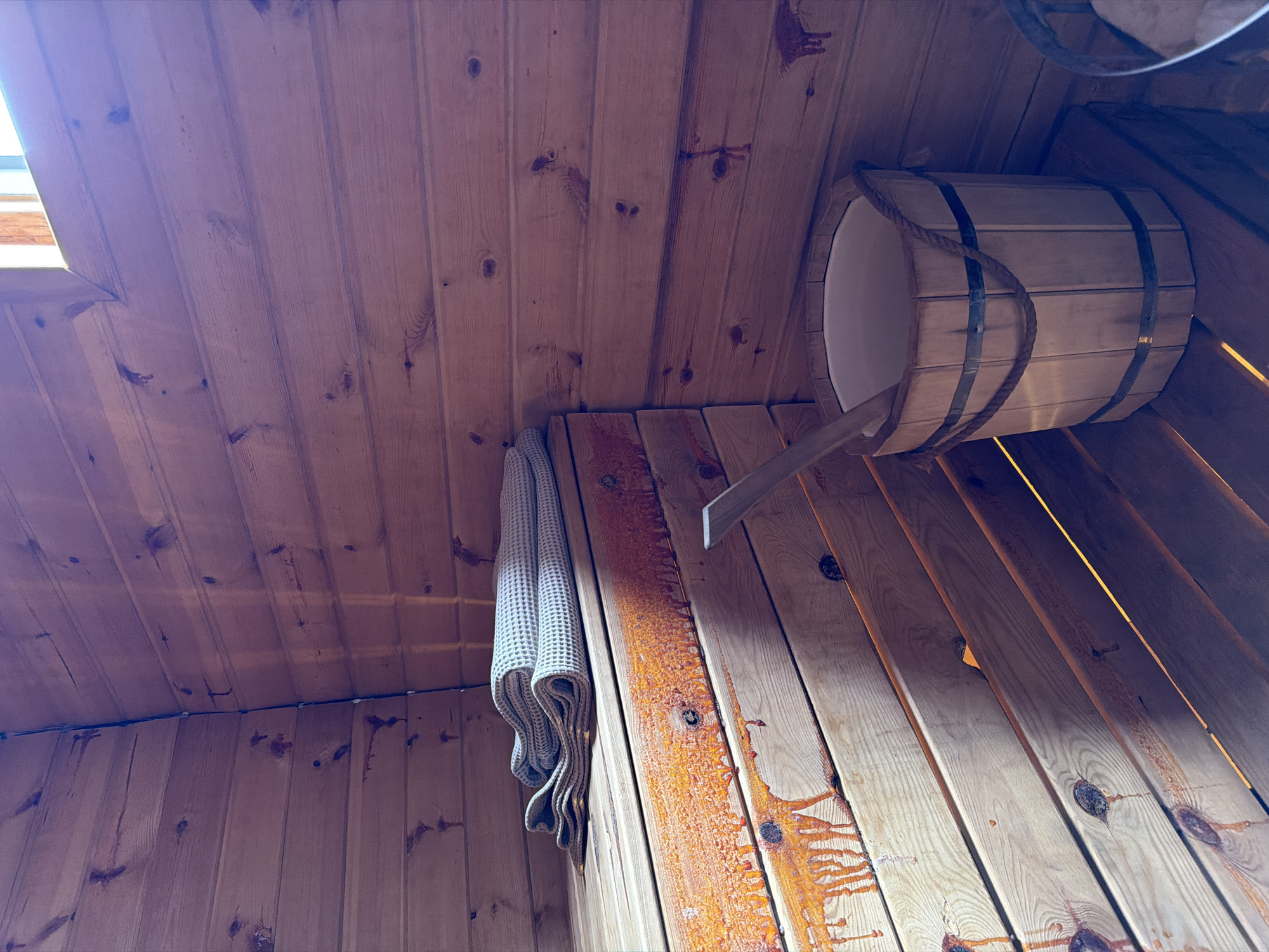 The image shows the interior of a wooden sauna. A corner of the room is visible, with walls and benches made of light-colored wood planks. Two folded towels, one lighter and one darker, are placed on the upper bench. To the right, a wooden bucket with a rope handle is positioned on a lower bench. A wooden ladle rests inside the bucket. The wood planks on the lower benches show dark, dripping stains, possibly from sap or resin. The scene is lit by natural light coming from an unseen window or opening at the top right of the frame. There are no people present in the image. The overall impression is one of a rustic and functional sauna environment.