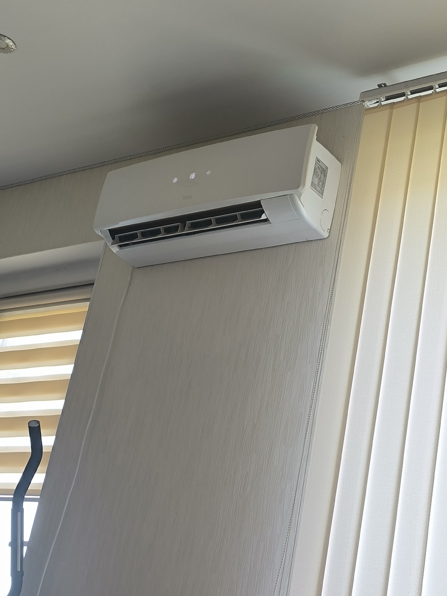 The image shows a white air conditioner unit mounted on a light-colored wall in an indoor setting. The air conditioner has a digital display showing "16" and some symbols. To the right of the air conditioner are vertical blinds, also in a light color. To the left, there are horizontal blinds. Part of a black and silver exercise equipment is visible in the lower left corner. The ceiling has a metallic trim. The image was taken in Chișinău, Moldova.