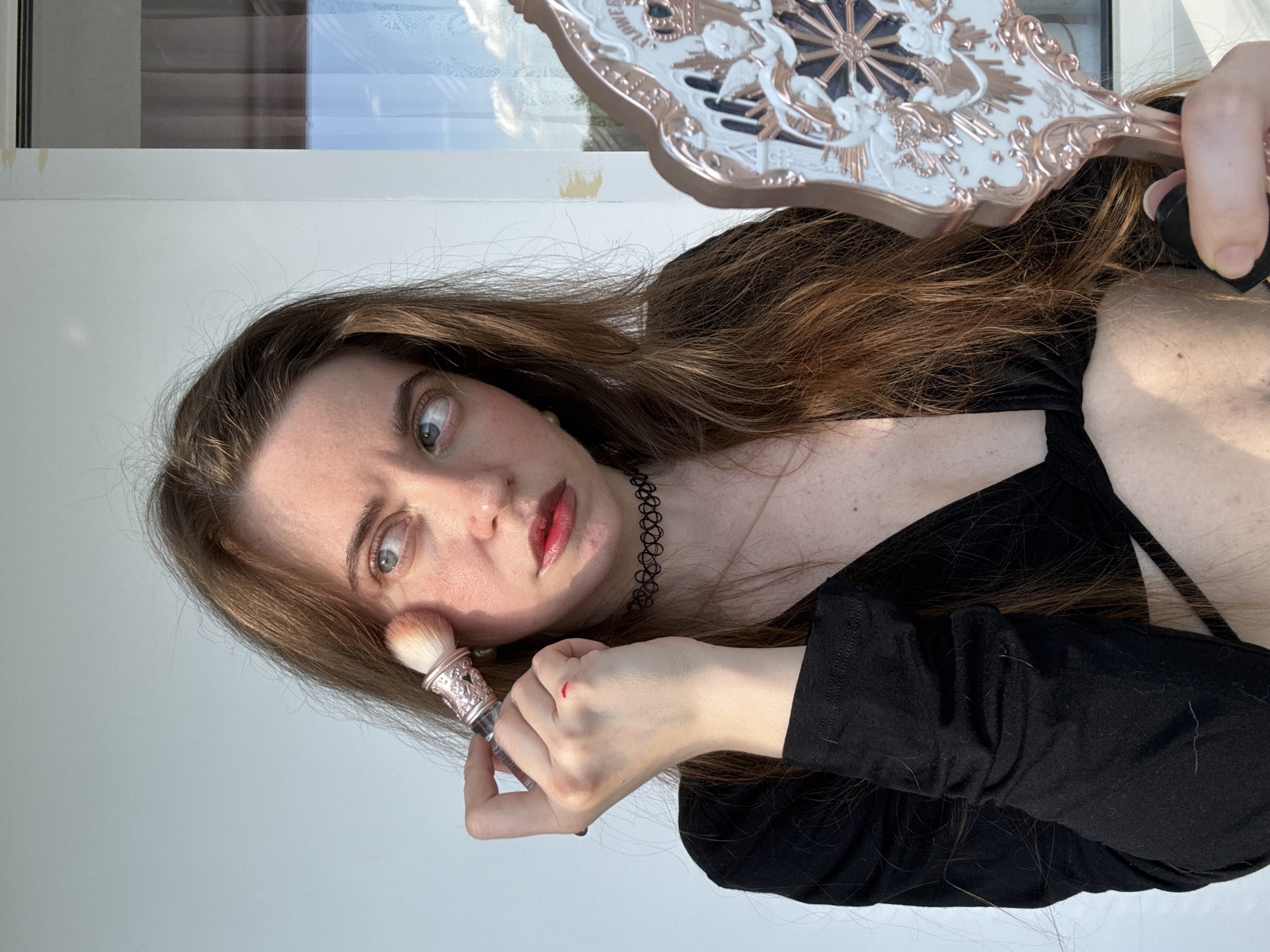 A young woman with long brown hair is applying makeup in front of a mirror. She is wearing a black top with a low neckline and a black choker necklace. Her left hand is holding a makeup brush to her cheek, and her right hand is holding a decorative mirror. The background appears to be an indoor setting with a white wall and a window showing a blue sky with clouds. There are no urban elements or landmarks visible to suggest a specific city location.
