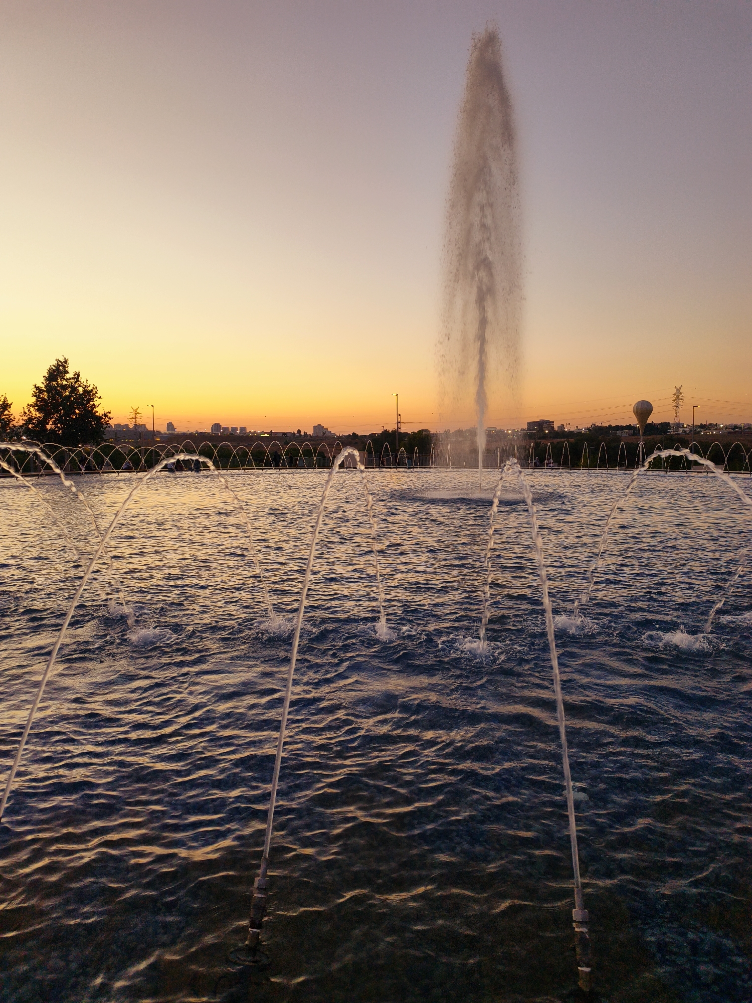 This image depicts a public fountain with multiple jets of water spraying upwards in a body of water, likely a pond or pool. In the foreground, several smaller water jets are visible, creating arcs of water. In the mid-ground, a larger, taller fountain is actively spraying a significant plume of water into the air. The water is disturbed by the jets, creating ripples and splashes. A decorative metal fence or railing with arched elements runs along the edge of the water body, separating it from the background.
The background features a tree line with several trees on the left side of the image. Beyond the trees, there is a cityscape with buildings and structures visible. A power line tower with a large balloon or object suspended from it is present in the mid-to-far background, along with other utility poles and infrastructure. The sky exhibits a gradient of colors indicative of a sunset or sunrise, with warmer yellow and orange tones near the horizon transitioning to a pale purple or pinkish hue higher up. The lighting suggests it is either early morning or late evening.
The location is identified as the fountain area in Be'er Sheva, Israel. The scene appears to be a public park or recreational area, with a serene and picturesque atmosphere due to the interplay of water, sky, and urban elements. No people are clearly discernible in the immediate foreground or mid-ground. The overall impression is a tranquil outdoor setting during twilight.