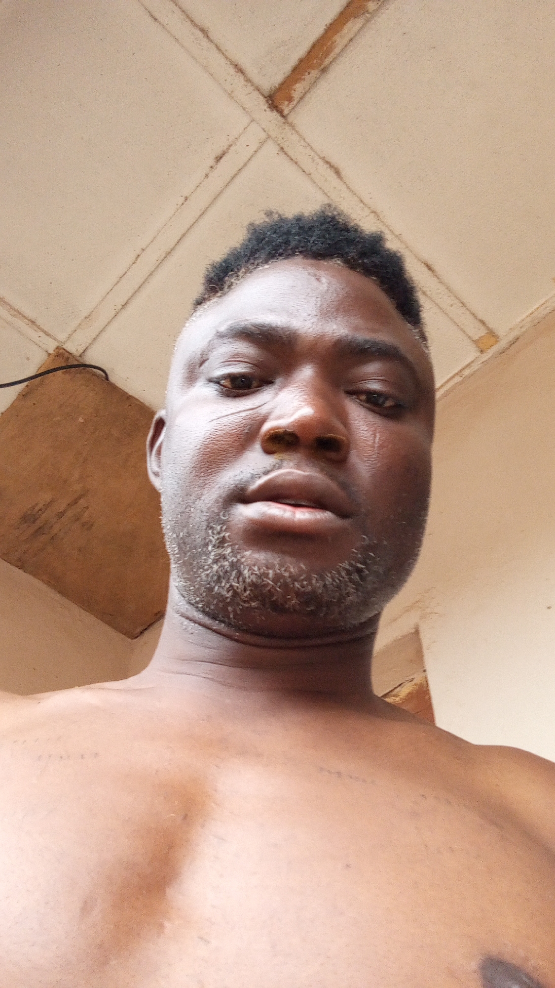 A close-up, low-angle selfie of a dark-skinned man with short, dark hair and a patchy beard. He has scars on his face. He appears shirtless. The background shows a light-colored ceiling with wooden beams and a section of a wooden board. The location is suspected to be Sabon Tasha, Nigeria.