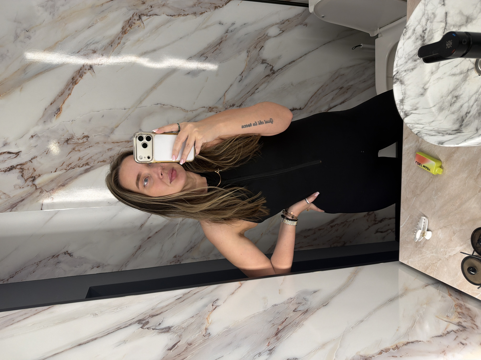 A woman with long, light brown hair is taking a mirror selfie in what appears to be a bathroom. She is wearing a black jumpsuit. The bathroom has marble-patterned walls and countertops, a white toilet, and a modern sink with a black faucet. There is a yellow object, possibly a bottle, and a pair of sunglasses on the countertop. The woman is holding a white iPhone. The location is not possible to determine from this image.