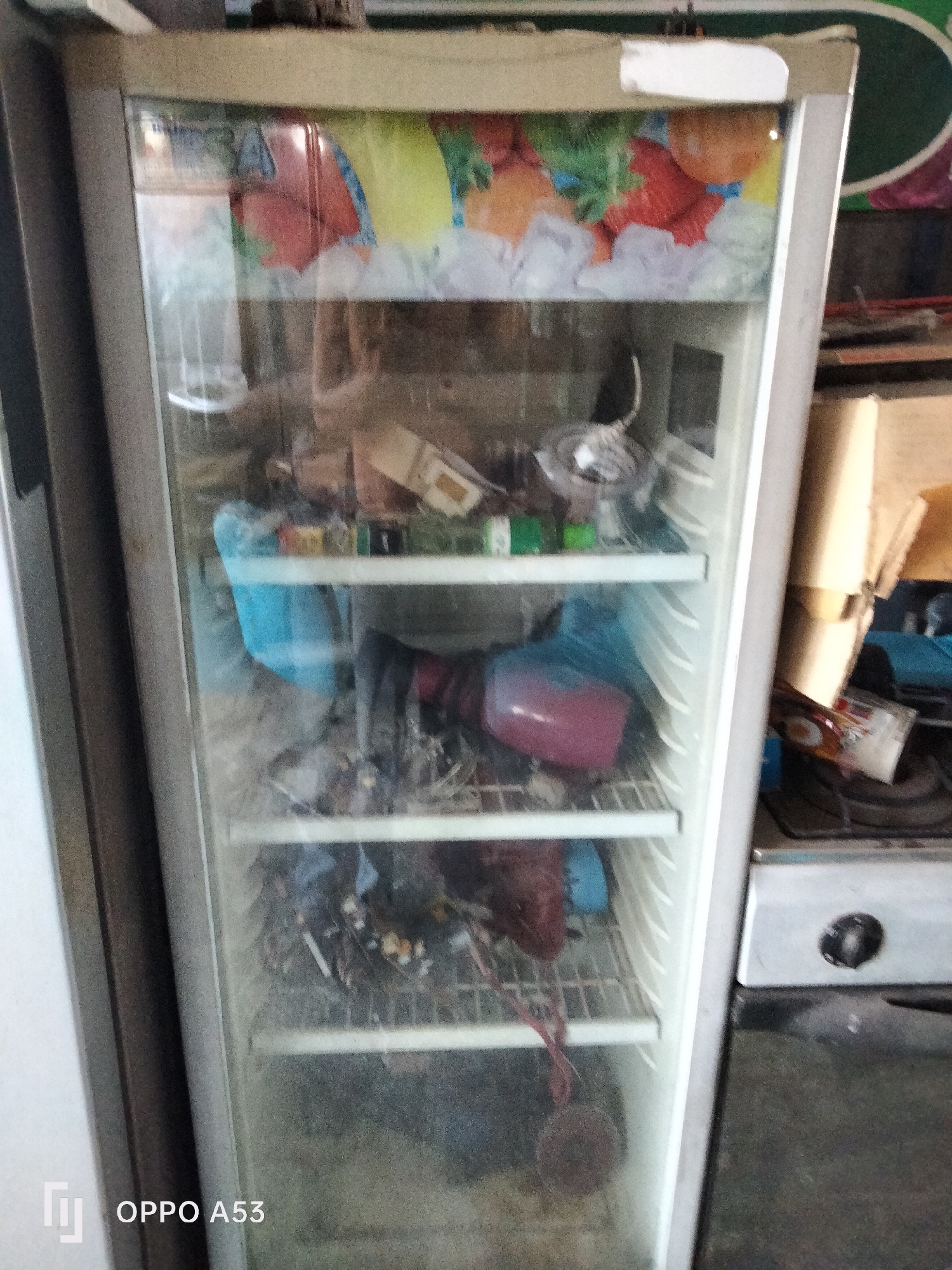 The image shows a commercial display refrigerator with its glass door slightly open. The refrigerator is filled with various items, including what appear to be bags of food, bottles, and possibly some frozen products. The interior is dimly lit, with shelves organized throughout. The exterior of the refrigerator is a light gray color, and the top section features a colorful advertisement depicting various fruits and ice. The overall setting suggests a retail or food service establishment. The image is captured from a slightly low angle, with the bottom right corner displaying "OPPO A53," indicating the device used for the photograph. The background is blurred, but suggests a cluttered environment with shelves and appliances. The scene is in Denpasar, Indonesia.