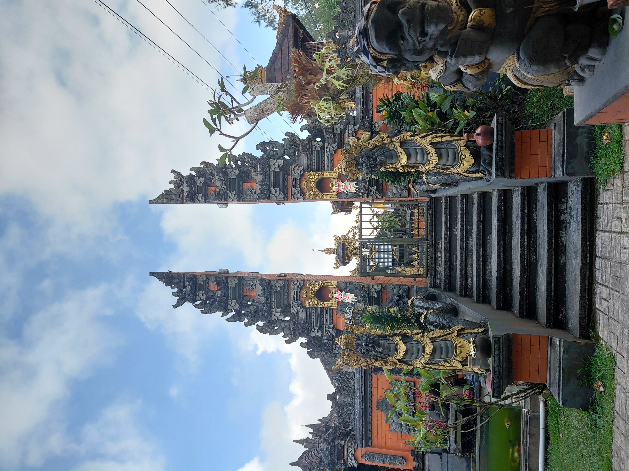 The image depicts the entrance to a Balinese temple in Sukasada, Indonesia, under a partly cloudy sky. The foreground is dominated by a set of stone stairs leading up to an ornate gateway. Flanking the stairs are two large stone statues of guardian figures, one on the left appearing serpentine and the other on the right resembling a demon. These statues are adorned with gold detailing. The gateway itself is an imposing structure with intricate carvings and decorative elements, featuring orange brickwork and darker stone accents. Beyond the gateway, a glimpse of the temple's architecture is visible, including a tiled roof and more decorative stonework. Lush green vegetation surrounds the entrance, including potted plants and trees. A small pond or water feature is visible to the left of the stairs, with what appear to be koi fish swimming within. Power lines are strung across the upper right portion of the image. The overall scene conveys a sense of traditional Balinese artistry and spiritual architecture.