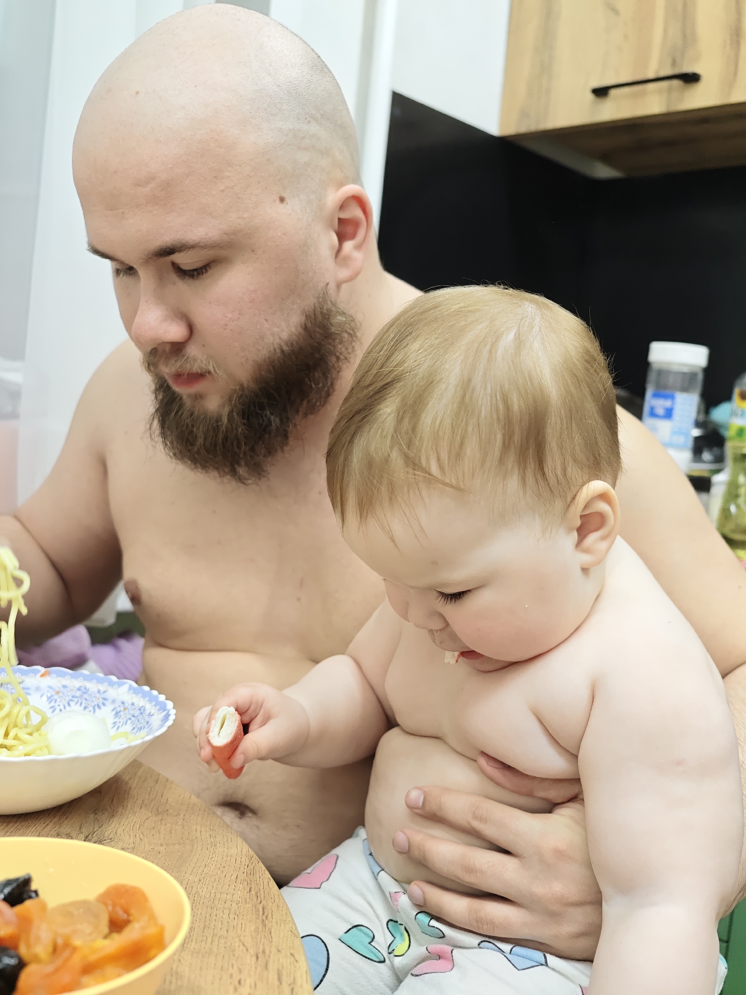 A bald, bearded man sits shirtless at a table with a baby on his lap. The baby, wearing patterned pajamas, holds a piece of food, likely a crab stick. The man is looking down at the table where there is a blue and white bowl of noodles and a yellow bowl of dried fruit. The scene appears to be in a kitchen setting with cabinets visible in the background. The location is Chaikovsky, Russia.