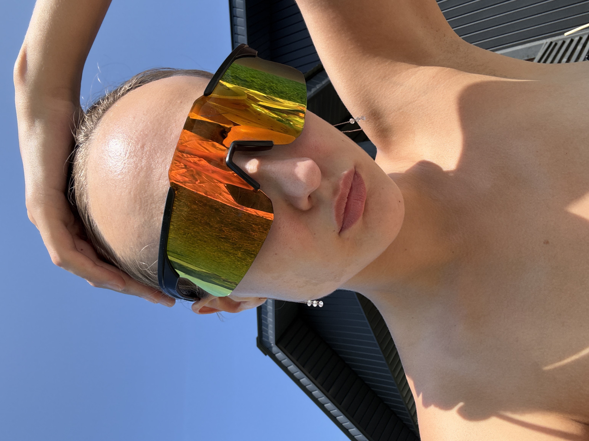 A young woman is captured from the chest up, against a clear blue sky and the side of a dark-paneled building, possibly in the city of Подшивалово, Russia. She wears large, mirrored sunglasses that reflect a vibrant orange and green landscape, suggesting an outdoor setting. Her right hand is raised, touching the top of her head, and a delicate earring with three small beads hangs from her left ear. Her skin is lightly tanned and appears to be dewy, possibly from the sun. The lighting suggests it is daytime.