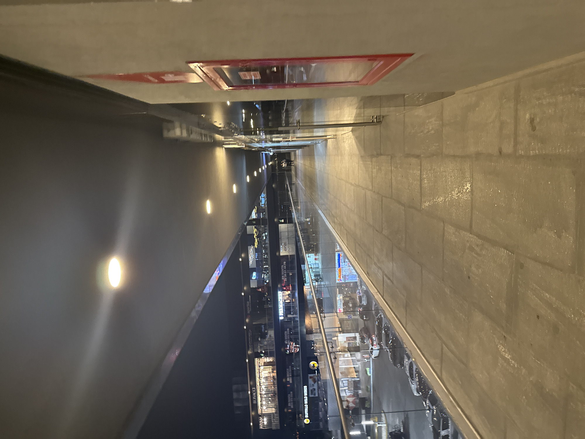 The image depicts a vertically oriented architectural scene, appearing to be an interior hallway with a view of a cityscape. The hallway is defined by a dark, reflective wall on the left and a textured, light-colored brick or stone wall on the right. Recessed lighting is visible along the dark wall. Overhead, there is a red-framed access panel. Between the two walls, a glass railing provides a view of an urban scene at night. Buildings with illuminated signs and advertisements are visible across the glass. In the reflected cityscape, businesses can be seen, along with street lights, vehicles, and pedestrians. The scene suggests a location overlooking a city center at night, possibly within a shopping mall or high-rise building. The location is San Bernardino Tlaxcalancingo, Mexico.