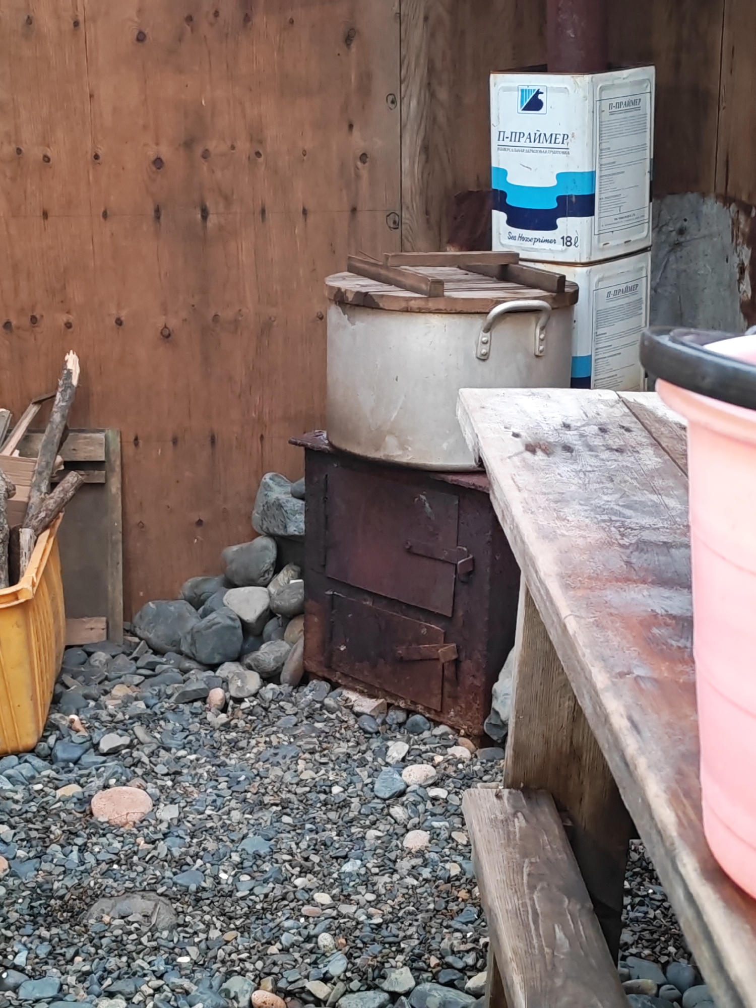 This is an outdoor scene in Reineke, Russia, featuring a rustic, weathered wooden structure serving as a backdrop. In the foreground, a wooden table made of rough planks is positioned at an angle, with visible nails and an aged patina. To the left of the table, a large, metallic pot with two handles rests on top of a dark metal stove, which appears to be a wood-burning type with two doors. The area around the stove and pot is covered with numerous smooth, grey and brown stones.
Behind the stove and pot, several large rocks are piled. To the far left, a yellow plastic container is filled with branches and pieces of wood. Further back, two large, white rectangular containers are stacked, each displaying blue and white labeling with Cyrillic text that appears to be "П-ПРАЙМЕР" (P-PRIMER) and "18 л" (18 liters). The wall of the wooden structure is made of plywood, showing visible grain and circular knots. The overall impression is one of a utilitarian outdoor space, possibly a backyard or a worksite, characterized by a mix of functional objects and natural elements.