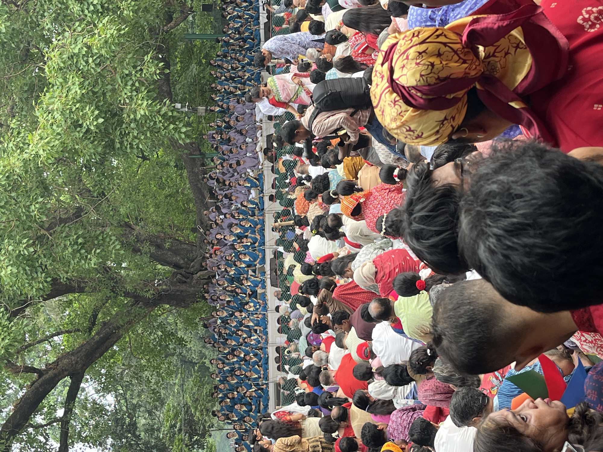 A large gathering of people, primarily of South Asian descent, are kneeling in a field at Suhrawardy Udyan, Dhaka, Bangladesh. A group of people wearing matching blue clothing sit apart from the rest of the crowd on the left side of the image. People in the crowd are wearing traditional South Asian clothing, including sarees and turbans. A large tree is visible on the left of the scene, providing shade over a segment of the crowd. The photograph appears to have been taken from an elevated perspective.