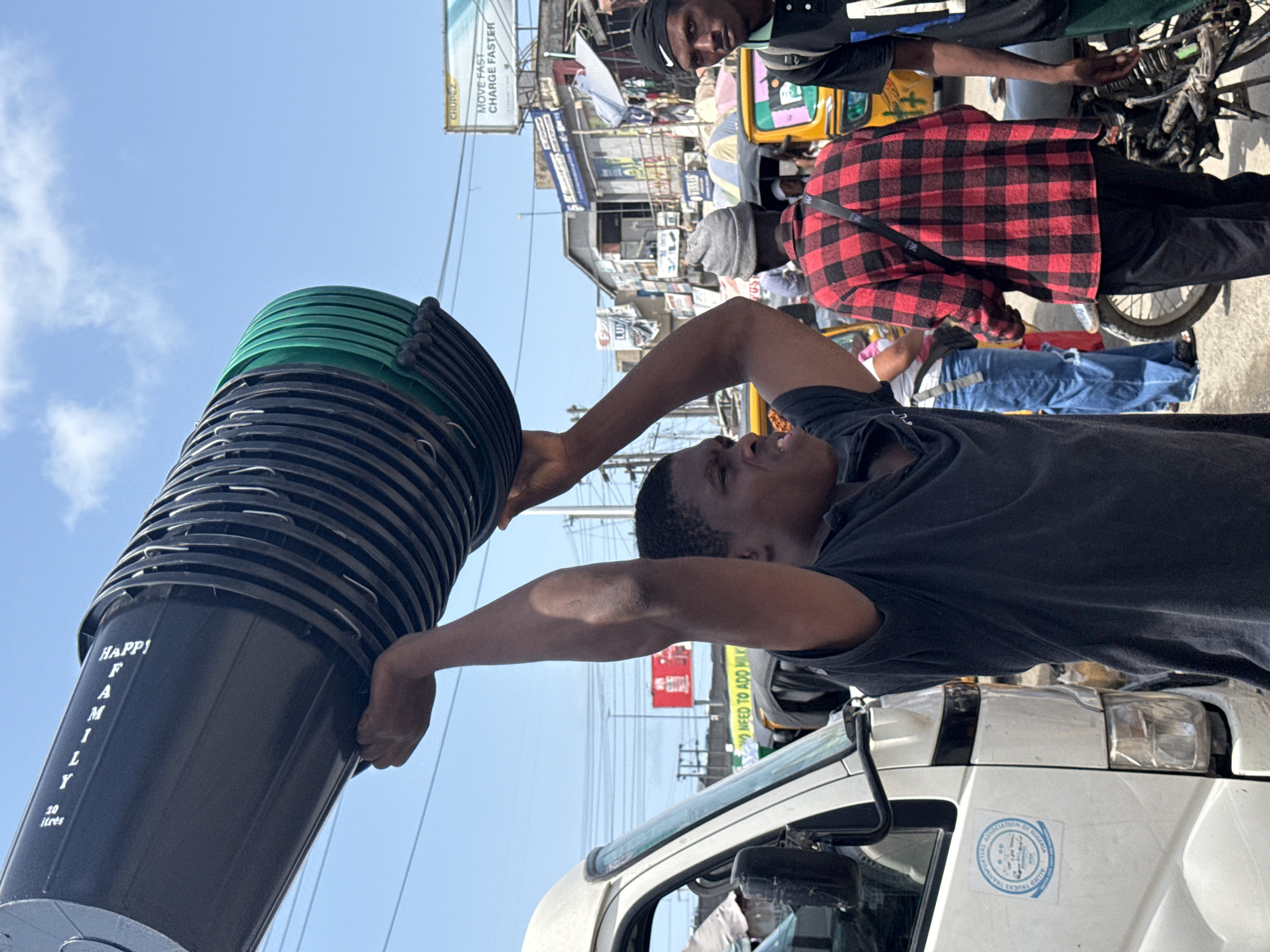 A man lifts a stack of black and green plastic buckets labeled "HAPPY FAMILY" over the roof of a white vehicle marked with the "ALLIED TRUCKS TRANSPORTERS ASSOCIATION OF NIGERIA" seal. The location appears to be a busy street in Ado, Nigeria. Other individuals are nearby, including one in a red and black plaid shirt, a man in a black shirt with a black hat, and people on bicycles and auto rickshaws. Advertisements and signs are visible in the background, including one advertising "MOVE FAST CHARGE FASTER".