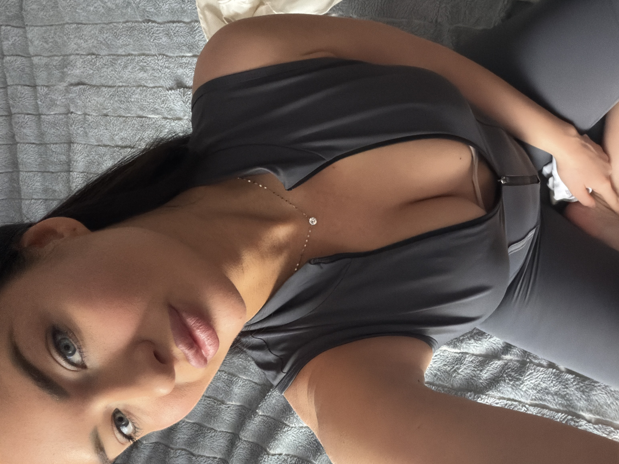 A woman is lying down on a gray blanket. She is wearing a gray outfit with a low-cut neckline. She is looking at the camera. A necklace is visible. No city or location context is provided in the image.