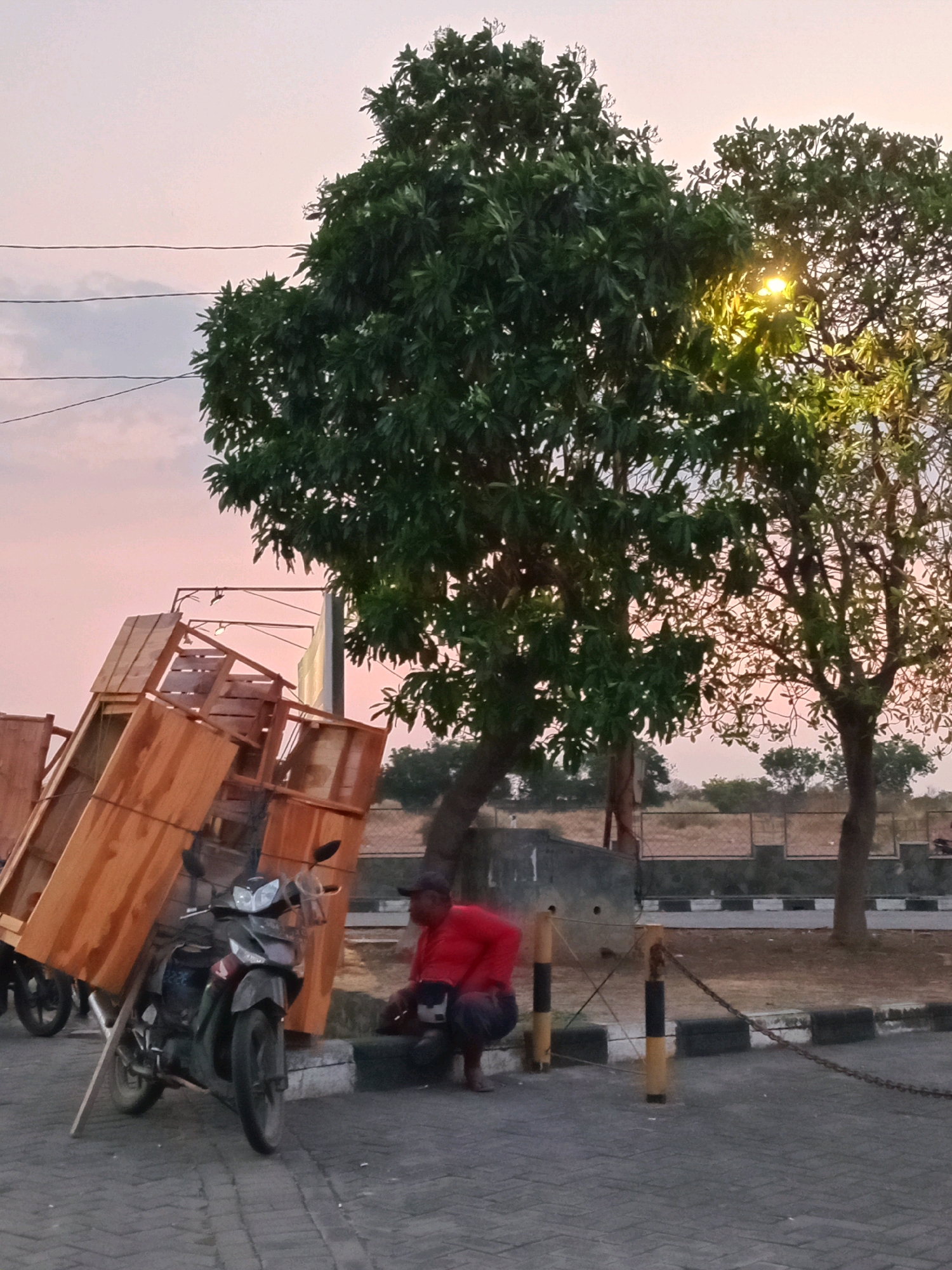 A motorcycle is parked on a paved surface, leaning against a wooden structure that appears to be a cart or trailer, loaded with planks of wood. A person wearing a red shirt and dark pants is seated on a concrete block next to the motorcycle, their body angled towards the wooden structure. In the background, several trees are visible, with the sky appearing to be a soft pink and orange, suggesting either sunrise or sunset. A street lamp is illuminated in one of the trees. The environment suggests an outdoor area, possibly a roadside or a parking area, with a fence and dry grass visible further in the background. The scene is located in Yosowilangun, Indonesia.