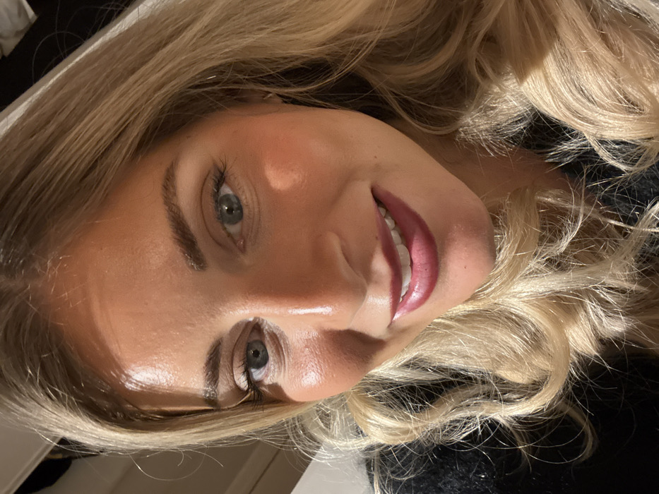 A woman with blonde wavy hair smiles at the camera. She has fair skin, light eyebrows, and blue-grey eyes. Her face is illuminated, reflecting light on her forehead and nose. She is wearing pink lipstick, which highlights her white teeth. The background includes a blurred black object. The image is rotated, with the woman's head tilted to the right.