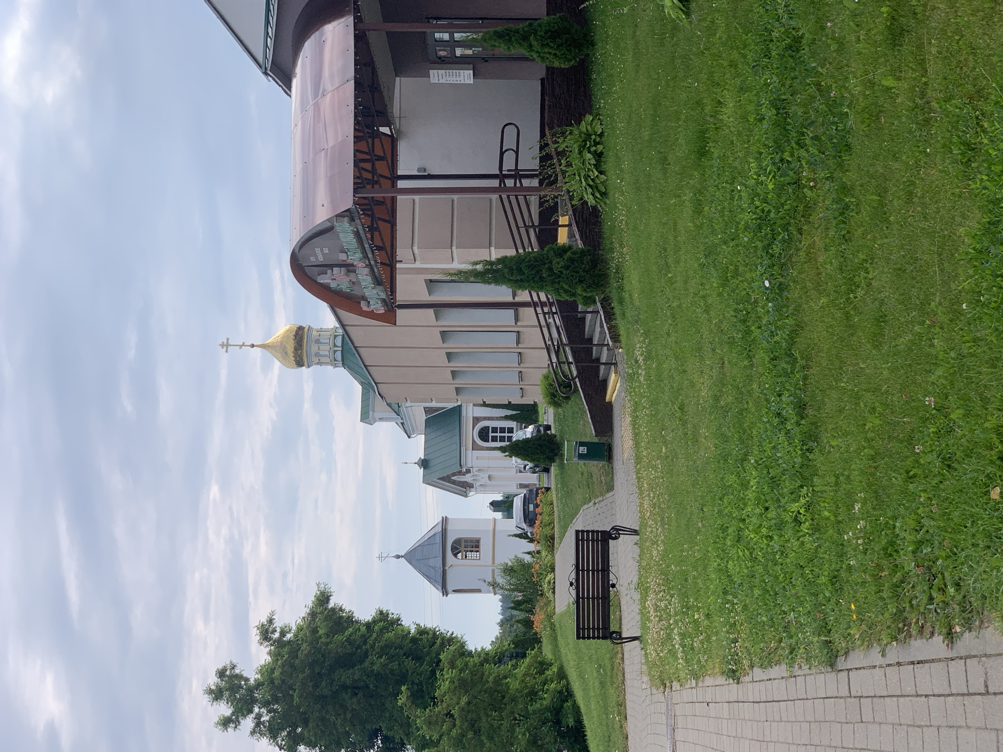 The image depicts a view of the Ascension Cathedral in Kapyĺ, Belarus. The foreground features a paved walkway with a brown wooden bench. To the right of the walkway is a well-maintained lawn with scattered small white flowers. Beyond the lawn, there is a building with a covered entrance featuring a translucent brown awning. Several evergreen trees are planted near the entrance.
In the mid-ground, the Ascension Cathedral is visible, characterized by its light-colored walls and a green roof topped with a golden dome and cross. Adjacent to the cathedral is a white bell tower with a grey conical roof and a cross. A black car is parked between the bell tower and another smaller building with a green roof.
The background shows a cluster of trees and a cloudy sky. The overall scene suggests a peaceful and well-kept religious complex in a temperate climate. There are no people visible in the image.
