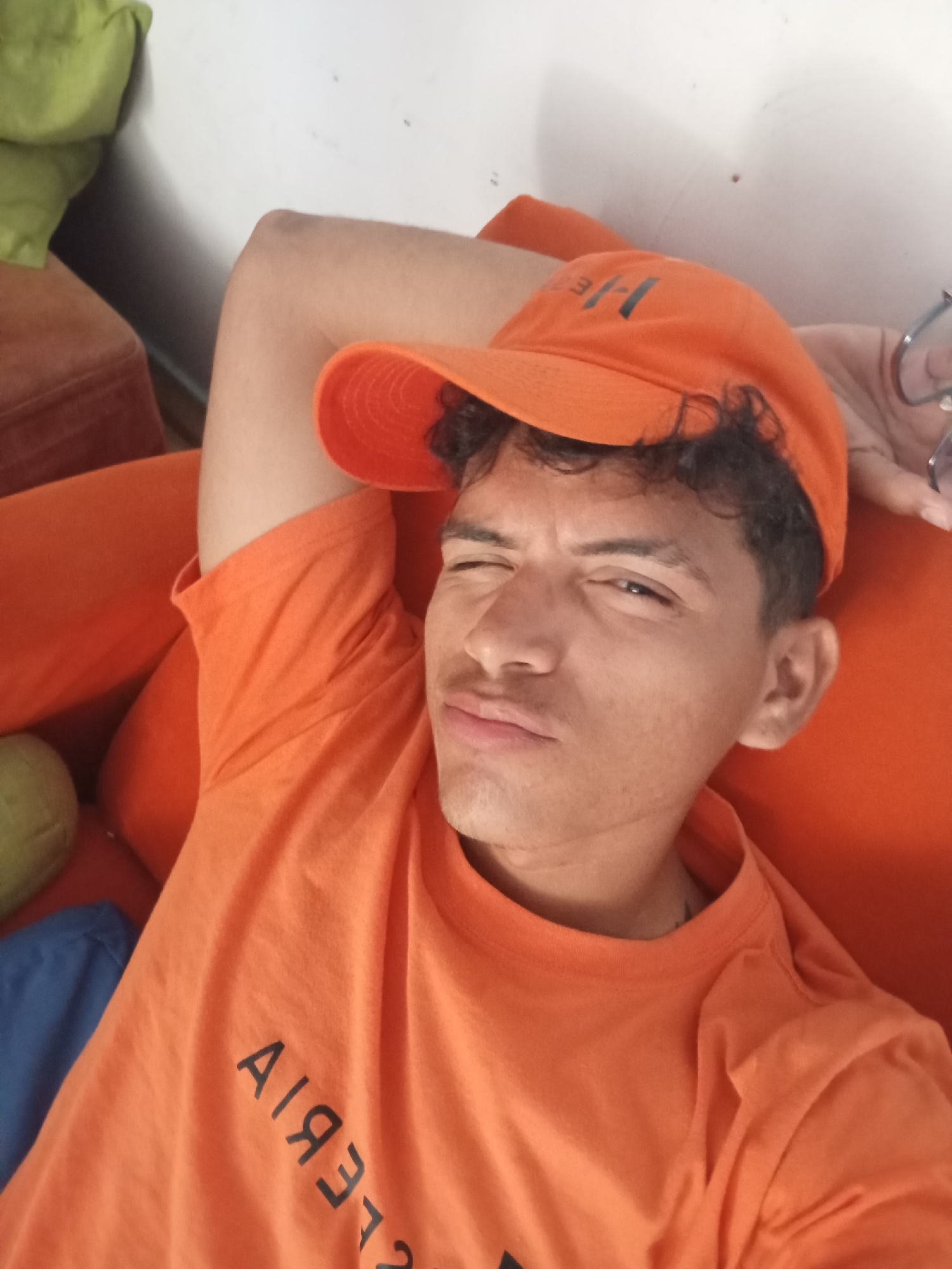 A man is featured in a selfie, reclining comfortably on an orange surface, likely a couch or bed. He wears an orange short-sleeved t-shirt and a matching orange baseball cap. The cap has the letters "H/E" visible on its front. His dark, curly hair is visible beneath the cap.
The man is looking towards the camera with a playful expression, winking or squinting his right eye and slightly puckering his lips. His left arm is bent behind his head, while his right hand, partially visible near his temple, holds a pair of glasses.
The orange t-shirt displays mirrored text on its chest, which appears as "AIЯЭTЭIA" in the image. The background consists of a plain white wall and parts of other furniture, including a light green cushion or pillow and a dark brown upholstered item. A blue object is also partially visible in the lower-left corner. The lighting suggests it is daytime. The overall scene conveys a relaxed and casual mood. The provided context indicates the location is Playa El Agua, Venezuela.