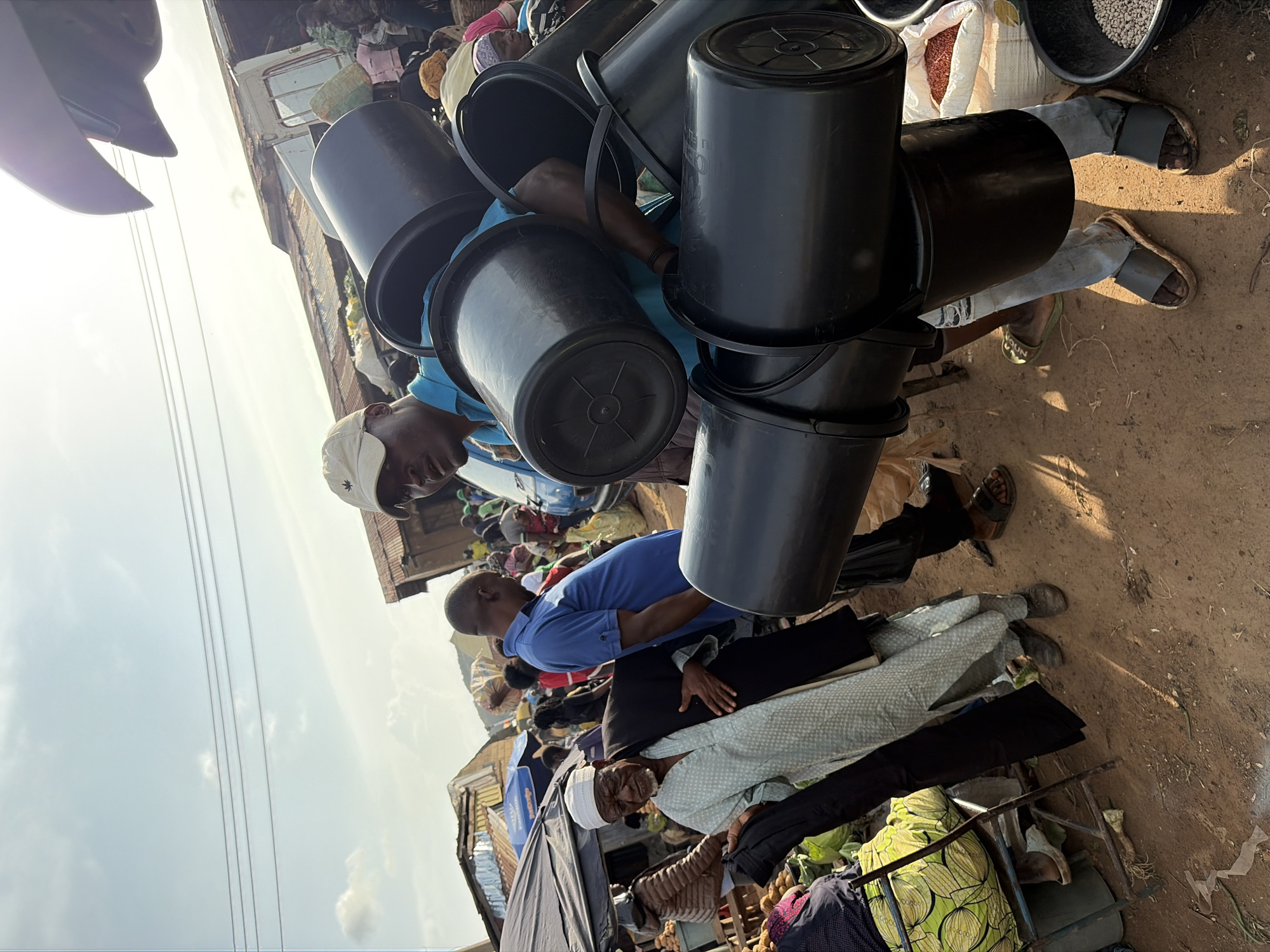 The image depicts a bustling outdoor market scene in Gura Top, Nigeria, under a bright, partially cloudy sky. In the foreground, a man wearing a blue shirt and a beige baseball cap is carrying a stack of large, black plastic buckets. His body is angled, and he appears to be in motion. To his left, an older man dressed in a light-colored patterned robe and a white cap is looking downwards, holding a dark garment in his hands. Behind them, other market-goers are visible, along with various stalls and structures, some with corrugated metal roofs, indicating a busy commercial area. The ground is dirt, with some scattered debris. In the background, a white van is parked, and tents or awnings provide shade. The overall impression is one of lively commerce in a typical Nigerian market setting.