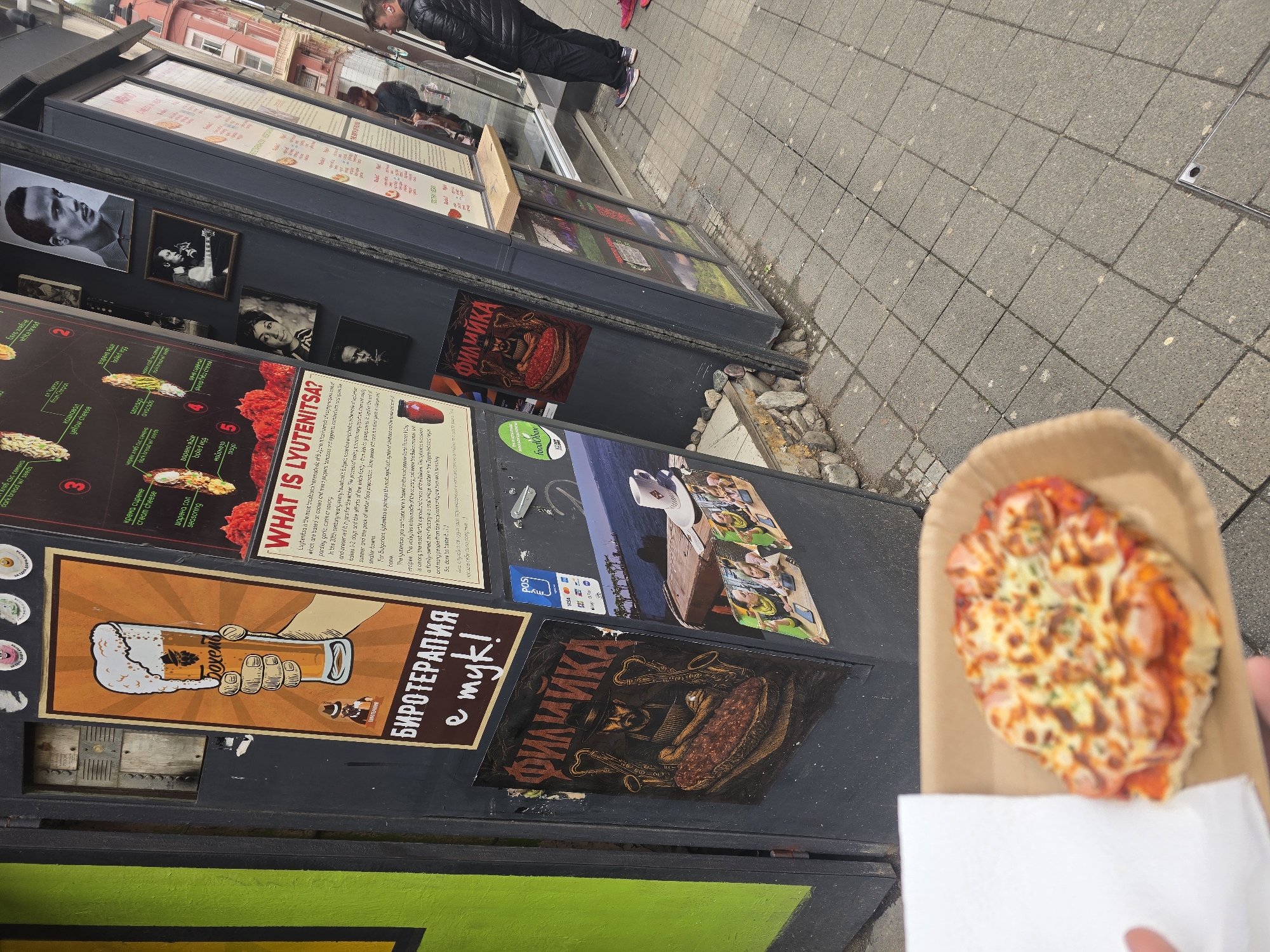 Here's a description of the image:
A close-up, slightly angled shot shows a small, round pizza on a brown cardboard tray, held in someone's hand with a white napkin. The pizza has a melted cheese topping with slices of sausage.
To the left, slightly out of focus, is a food stand, likely in Sofia, Bulgaria based on the presence of signs in Bulgarian. The stand has dark gray exterior panels covered in food advertisements and images. There is text reading "ФИЛИЙКА" in Cyrillic, and advertisements for a beer called "Бохемь." A poster is also visible describing a food item called "LYUTENITSA," a popular Bulgarian food. A sign indicates that VISA is accepted. A strip of bright green paint appears at the bottom.
In the background, on the right, is a sidewalk paved with gray stone tiles. Further back, a few people are walking on the sidewalk next to the food stalls. The overall composition shows a street-side food vending operation in an urban environment.