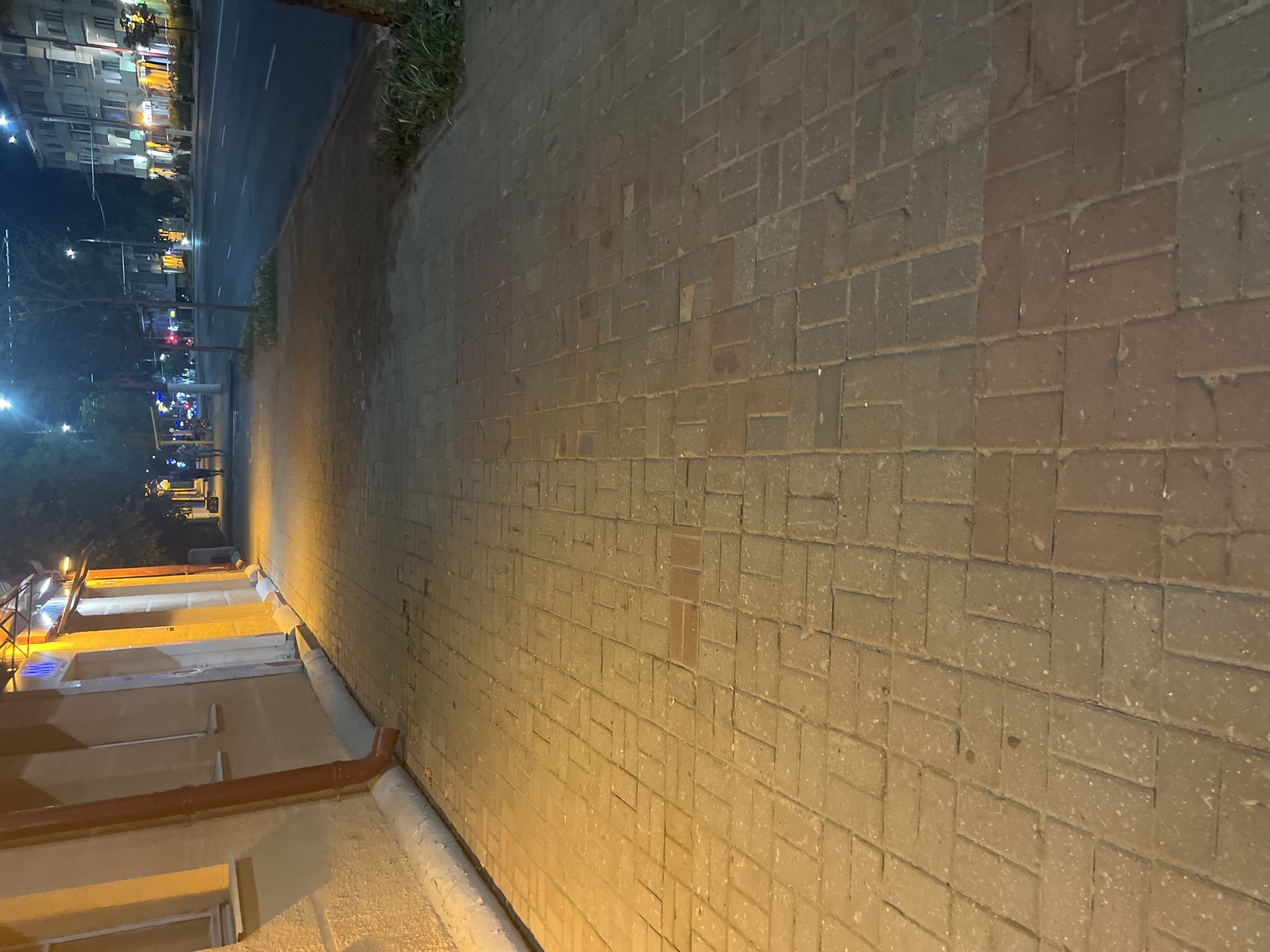 The image shows a brick sidewalk in the city of Vitsebsk, Belarus, at night. The sidewalk is illuminated by artificial light, casting a warm glow. To the left of the sidewalk, there is a building with a light-colored facade and a red downspout. Further down the sidewalk, streetlights and building lights are visible, illuminating the street and sidewalks on the opposite side. The street appears to be busy, with cars and people present. The overall scene depicts a typical urban streetscape at night.