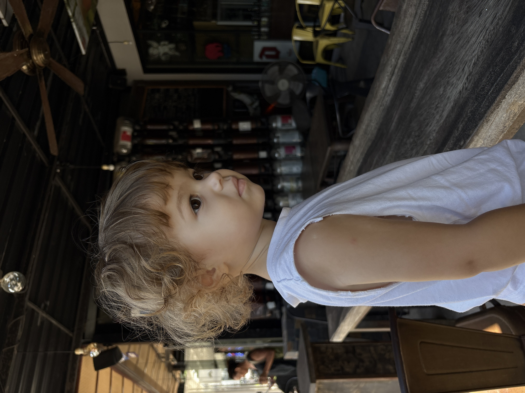 A young child with curly blonde hair and a white sleeveless shirt is pictured in profile, facing right. The child's right eye is visible. They are standing outdoors, in front of a wooden counter or bar. In the background, there are tables, chairs, a ceiling fan, and what appears to be a restaurant or bar setting. A disco ball is visible hanging from the ceiling. The location is identified as being in the city of Choeng Thale, Thailand.