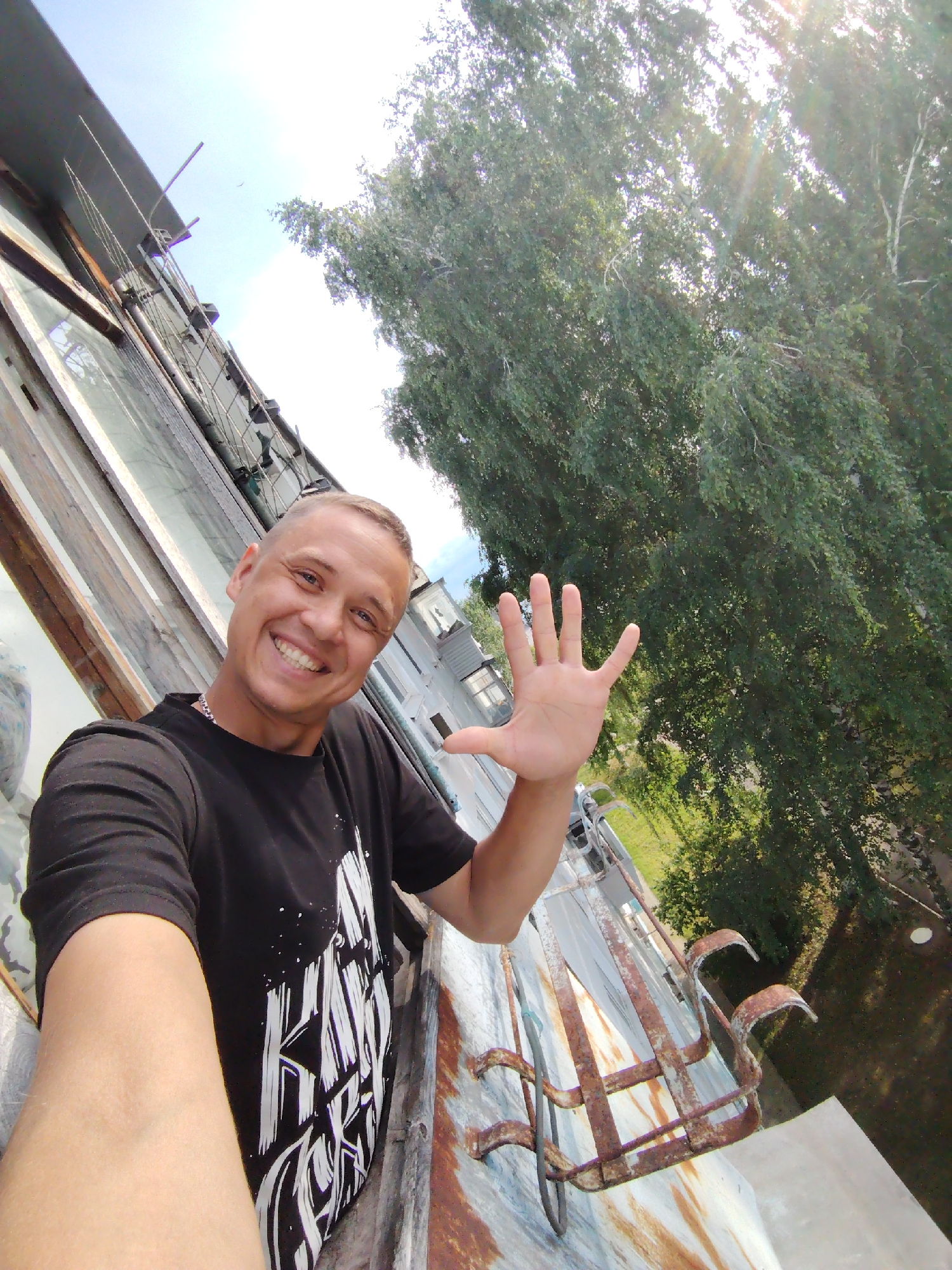 A man with a shaved head and a wide smile is taking a selfie from a balcony. He is wearing a black t-shirt with white text and graphics. His left hand is raised, palm facing the camera, with all five fingers extended. The man is standing on a balcony with a rusted metal railing. Behind him, to the left, is the side of a building with large windows. To the right and in the background, lush green trees fill the frame, with sunlight filtering through the leaves. The sky is bright blue and clear, suggesting it is daytime. The overall mood is cheerful and positive. The image appears to have been taken in Chaikovsky, Russia, given the provided location context. There is no visible text in the image other than on the man's t-shirt, which is partially obscured by his arm.