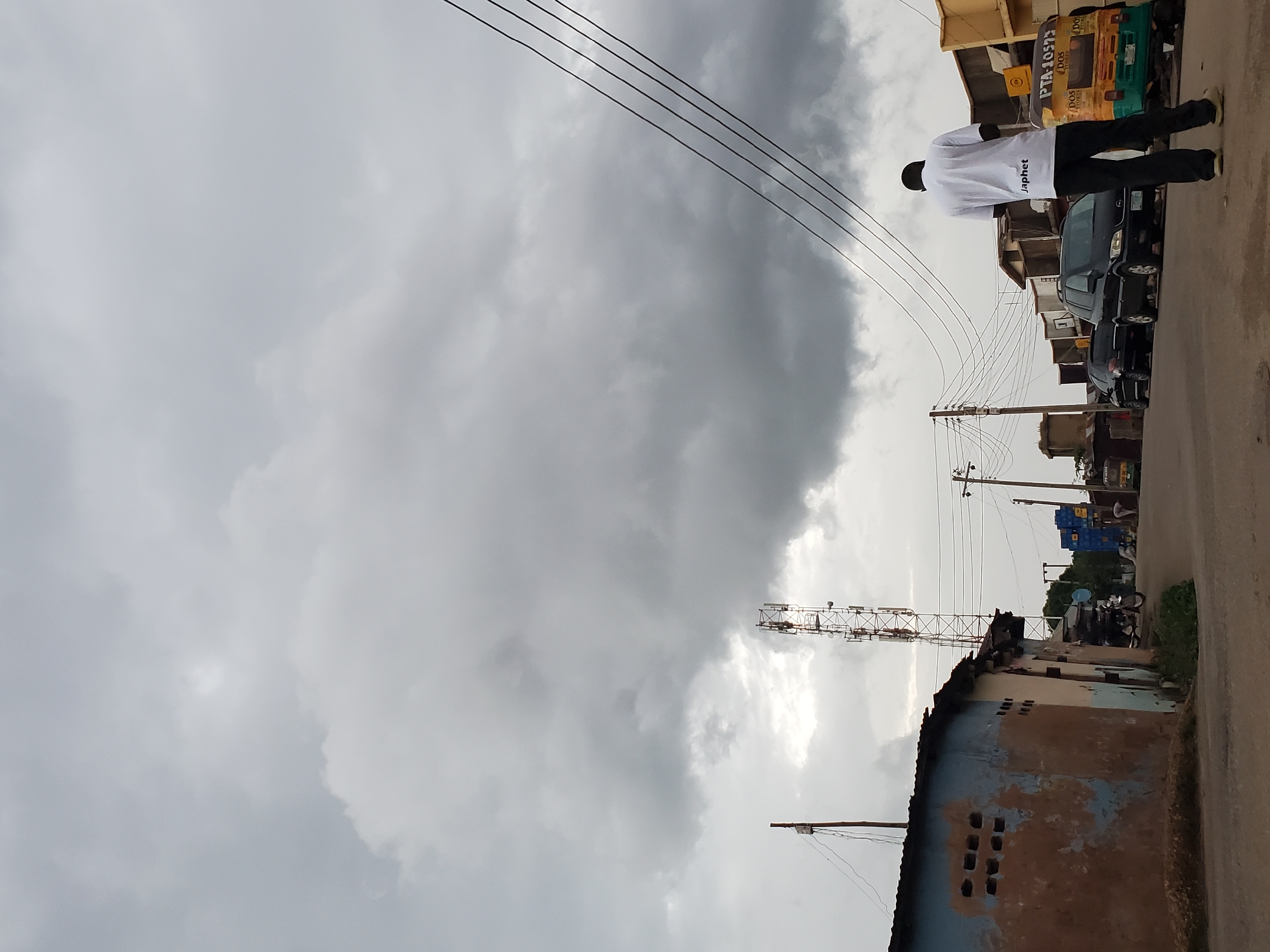In Jos, Nigeria, a man wearing a white t-shirt with "Japhet" printed on the back stands on a dusty road, facing away from the camera. The sky is overcast with dramatic, dark gray clouds. To the left of the frame, a dilapidated building with faded blue and brown paint features several rectangular openings. A tall telecommunications tower rises in the background, its metallic structure visible against the cloudy sky. Several cars are parked along the right side of the road, including a black SUV and what appears to be a three-wheeled transport vehicle with yellow and green markings. Power lines crisscross the sky, strung between utility poles. The overall scene depicts a somewhat neglected urban environment under a heavy sky.