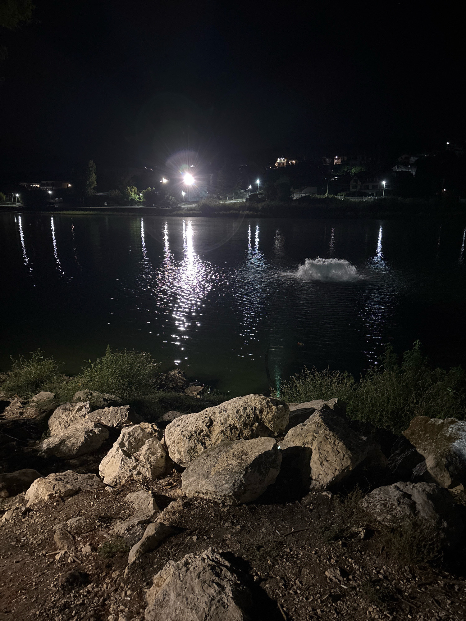 The image shows a nighttime view of a lake in Nikolovo, Bulgaria. The lake is illuminated by artificial lights, casting reflections on the water's surface. In the foreground, a rocky embankment is visible, with scattered stones and dry vegetation. On the lake, a fountain or aerator is active, creating a large plume of white water. In the background, the silhouette of a town or village can be seen with lights from buildings and street lamps scattered along the hillside. The overall atmosphere is tranquil and dark, characteristic of a rural or suburban area at night. There are no people or animals visible in the image.