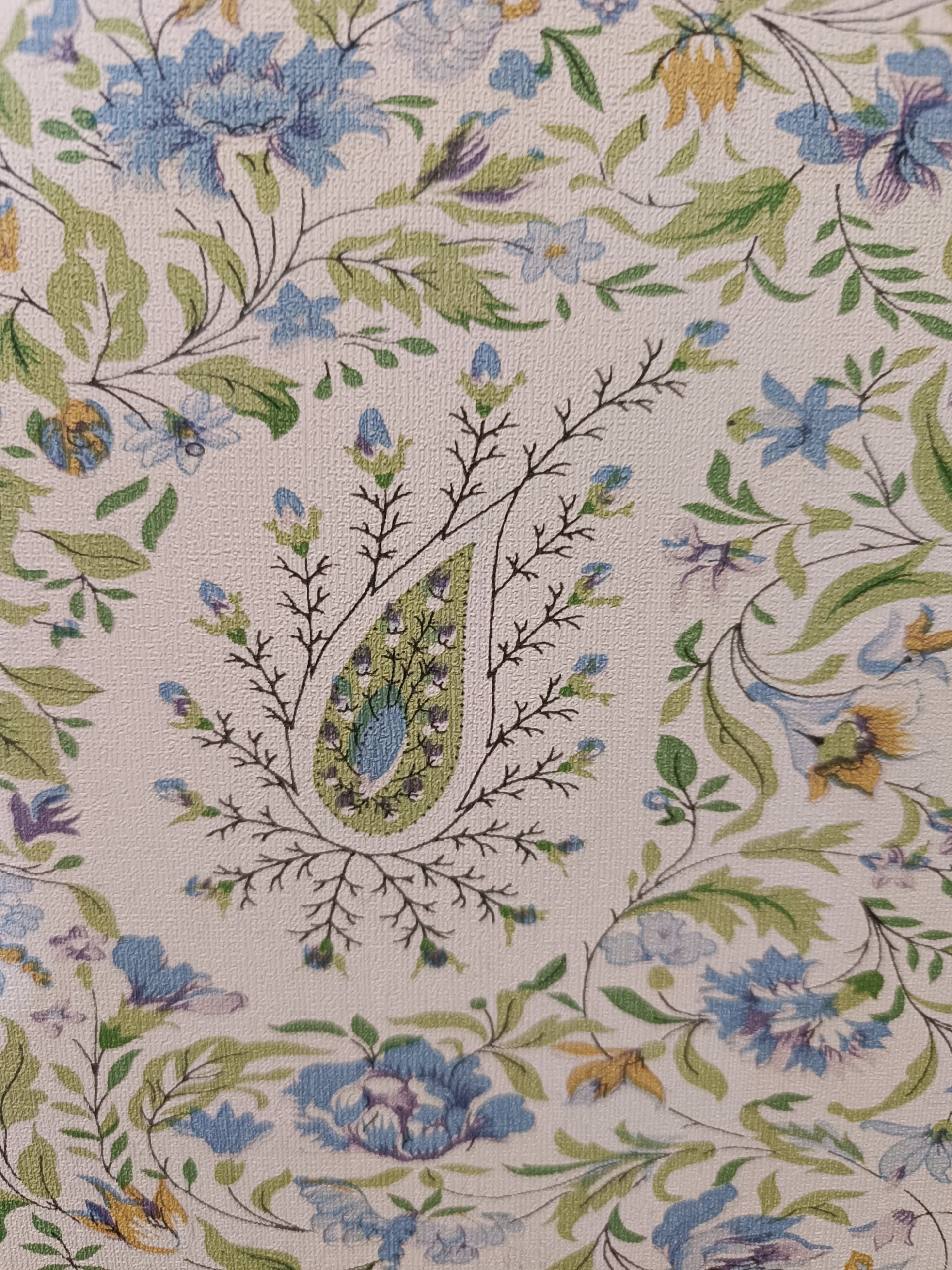 The image is a close-up of a fabric with a floral paisley pattern. The pattern features delicate blue and purple flowers, interspersed with green leaves and thin, dark brown branches. In the center, a prominent paisley motif is rendered in shades of green and blue. The fabric has a textured surface, possibly linen or cotton. The image is focused on the pattern itself, with no environmental context or people visible. The texture of the fabric suggests it could be a decorative cushion, upholstery, or fabric intended for clothing. The style of the pattern evokes traditional Indian textiles, particularly those from the Kashmir region, known for its intricate floral and paisley designs. The city of Srinagar is famous for its handicrafts, including exquisite pashmina shawls and carpets that often feature such detailed floral motifs. While the image doesn't depict Srinagar directly, the style of the pattern strongly aligns with the artistic heritage of the region.