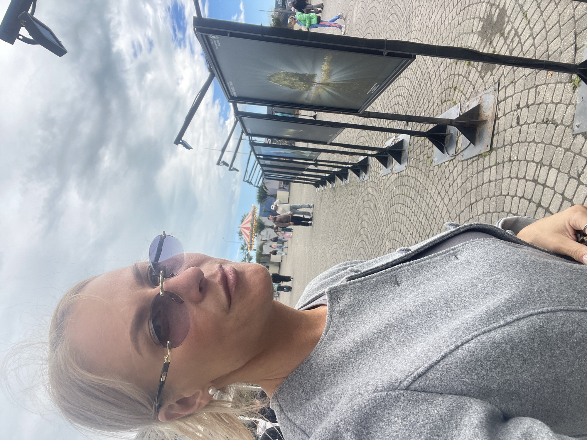 A street scene is depicted in a tilted, medium-shot selfie of a woman wearing sunglasses and a gray coat. The background features a row of outdoor photograph displays lining a paved area in the city of Sochi, Russia. Some people are standing near a distant carousel, visible in the background. The sky is overcast with gray and white clouds. The photo appears to be taken at ground level.