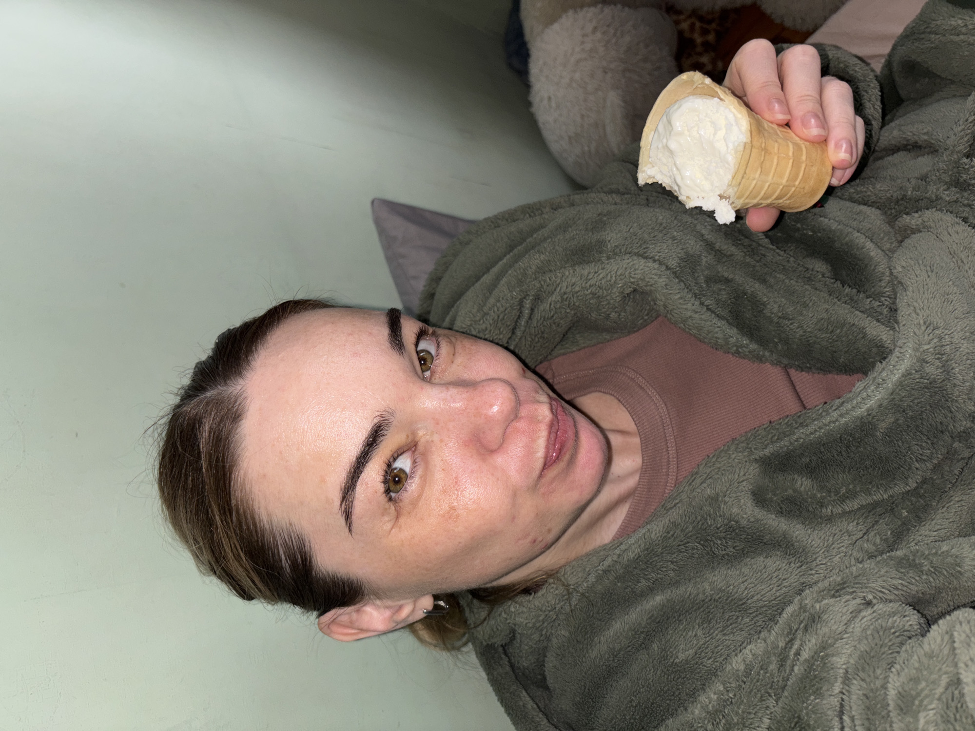A person is holding an ice cream cone with a bite taken out of it. The person is wearing a brown shirt and a green fuzzy jacket. The background is a light green wall. A pillow can be seen behind the person.