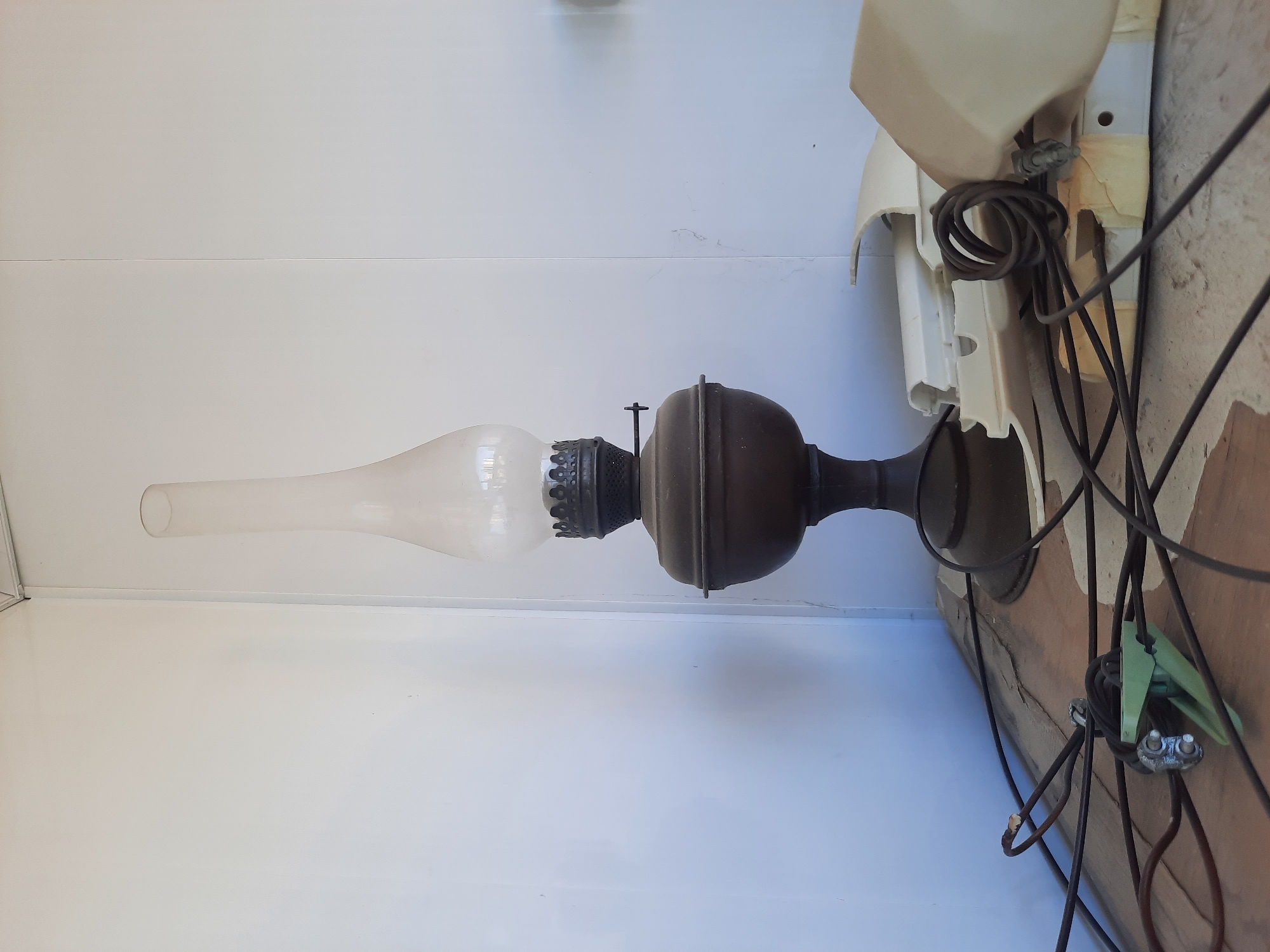 An antique oil lamp with a clear glass chimney stands on a wooden surface against a plain white wall. The lamp has a dark, metal base and a wide, rounded reservoir. To the right of the lamp, a pile of white plastic debris, possibly from a fan or appliance, is visible. Several black electrical wires are strewn across the wooden surface in the foreground, some tangled together, and one set is attached to a green clamp with metal fasteners. The environment appears to be indoors, in an area that is not well-lit.
