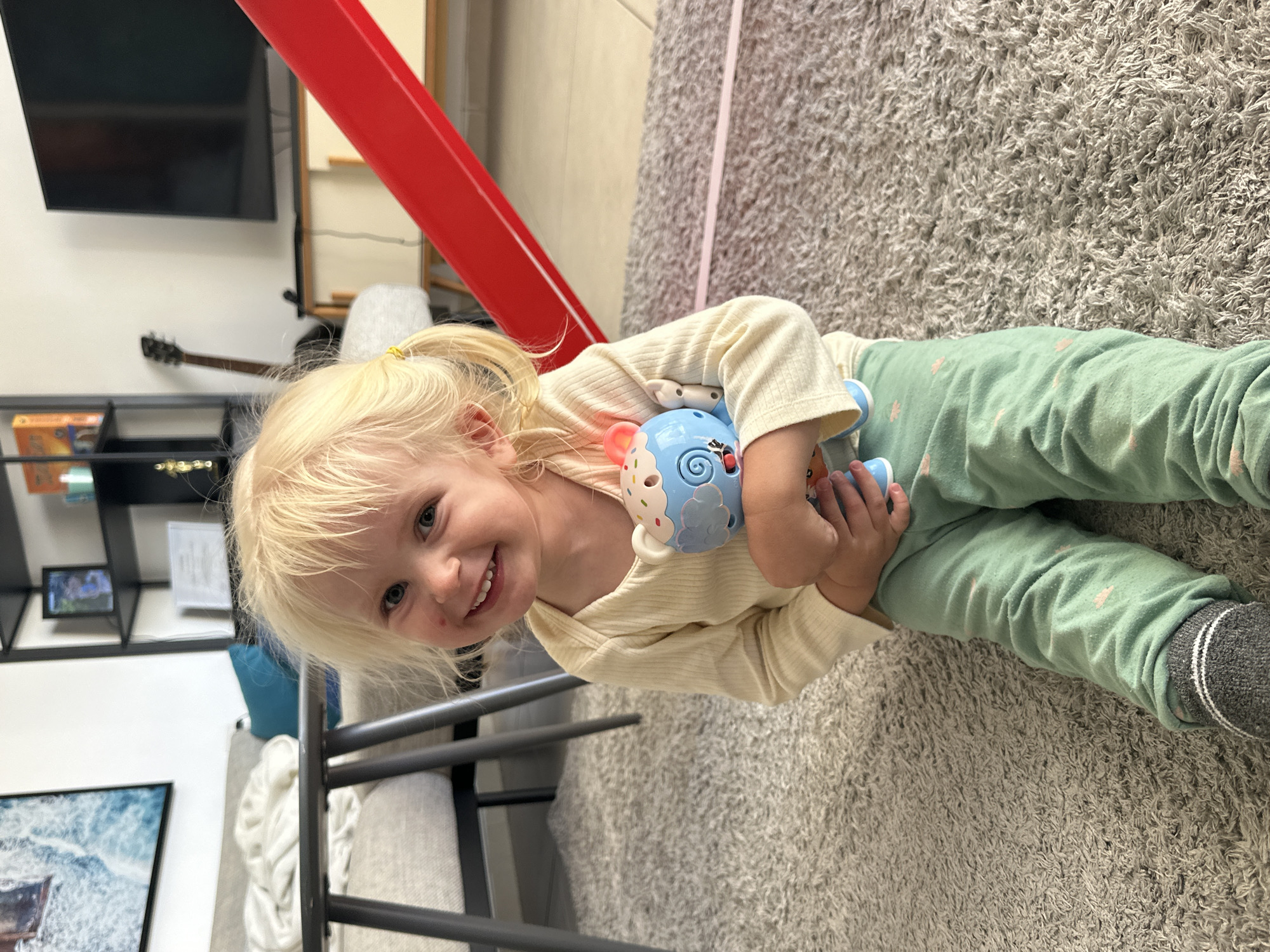A young Caucasian girl with blonde pigtails sits on a grey carpet, smiling at the camera. She is wearing a cream-colored long-sleeved shirt and light green pants with a pink star pattern. In her arms, she clutches a blue stuffed toy with a cupcake-like decoration on its head. The background shows a living room with a couch, a coffee table, shelves with books and decorations, a television, and a red structure that appears to be part of a play area. The scene is indoors, likely in a residential setting in Petah Tikva, Israel.