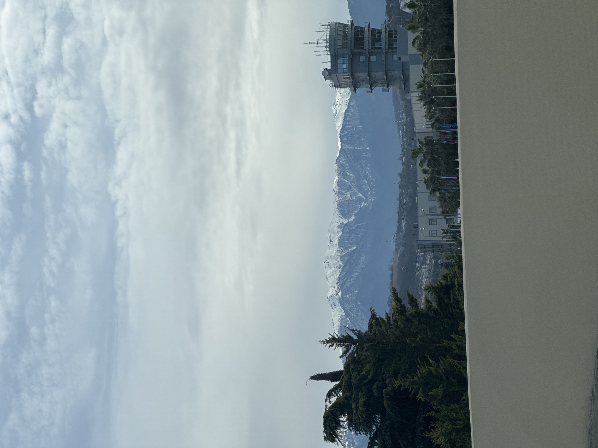 The image captures a view in Sochi, Russia, featuring a mountain range partially covered in snow, a multi-story building with antennae, palm trees, evergreen trees, other smaller buildings, and a cloudy sky. In the right foreground is a vertical section of a wall. Flags are shown in a row in the mid-ground.