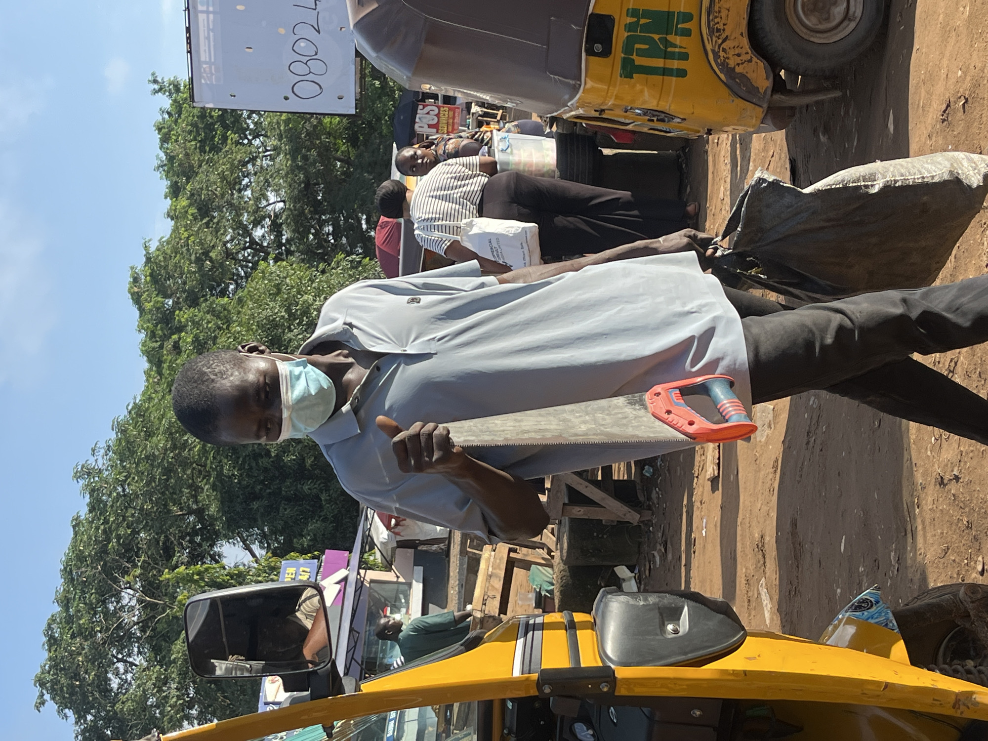 A man wearing a light blue polo shirt, dark pants, and a light blue face mask walks through a dusty outdoor market in Abuja, Nigeria. He is carrying a large hand saw with a red and blue handle in his right hand and a dark-colored plastic bag in his left hand. The man is positioned in the foreground, facing towards the right of the frame. In the background, a yellow auto-rickshaw is visible on the left, and another yellow auto-rickshaw with the letters "TPN" painted on its side is on the right. Other people and market stalls are also present in the background, along with trees and a bright blue sky. A white sign with black writing, including the number "08024", is visible above the market stalls on the right.