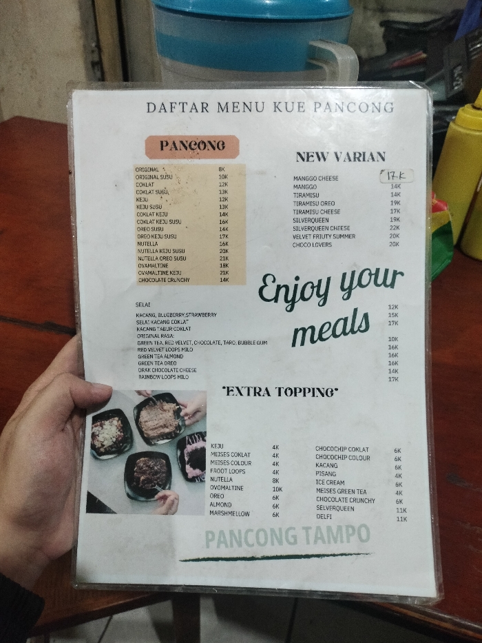 A hand holds up a laminated menu for "Kue Pancong" (Pancong Cake), likely in an indoor eatery in Dramaga, Indonesia. The menu, titled "DAFTAR MENU KUE PANCONG," details various offerings and prices, indicated in thousands of Rupiah ("K").
The main "PANCONG" section lists flavors such as Original, Susu, Coklat, Keju, Oreo, Nutella, Ovomaltine, and Chocolate Crunchy, ranging from 8K to 21K. A "NEW VARIAN" section features options like Manggo Cheese, Tiramisu, Silverqueen, Velvet Fruity Summer, and Choco Lovers, priced between 14K and 22K.
Additional categories include "SELAI" (Jams) with Kacang, Blueberry Strawberry (12K), Kacang Coklat (15K), and Kacang Tabur Coklat (17K). "ORIGINAL RASA" lists flavors such as Green Tea, Red Velvet, Chocolate, Taro, Bubble Gum (10K), Red Velvet Loops Milo (16K), Green Tea Almond (16K), Green Tea Oreo (16K), Drak Chocolate Cheese (14K), and Rainbow Loops Milo (17K).
An "EXTRA TOPPING" section provides a wide array of additions, including Keju, Meises Coklat/Colour, Froot Loops, Nutella, Ovomaltine, Oreo, Almond, Marshmellow, Chocochip Coklat/Colour, Kacang, Pisang, Ice Cream, Meises Green Tea, Chocolate Crunchy, Silverqueen, and Delfi, priced from 4K to 11K. The decorative text "Enjoy your meals" is prominently displayed, and "PANCONG TAMPO" is visible at the bottom, likely indicating the shop or brand.
The menu displays images of several prepared Pancung cakes in square dishes. In the blurry background, a blue container and yellow squeeze bottles can be seen on a dark surface. The lighting appears dim, suggesting an indoor setting, possibly during the evening.