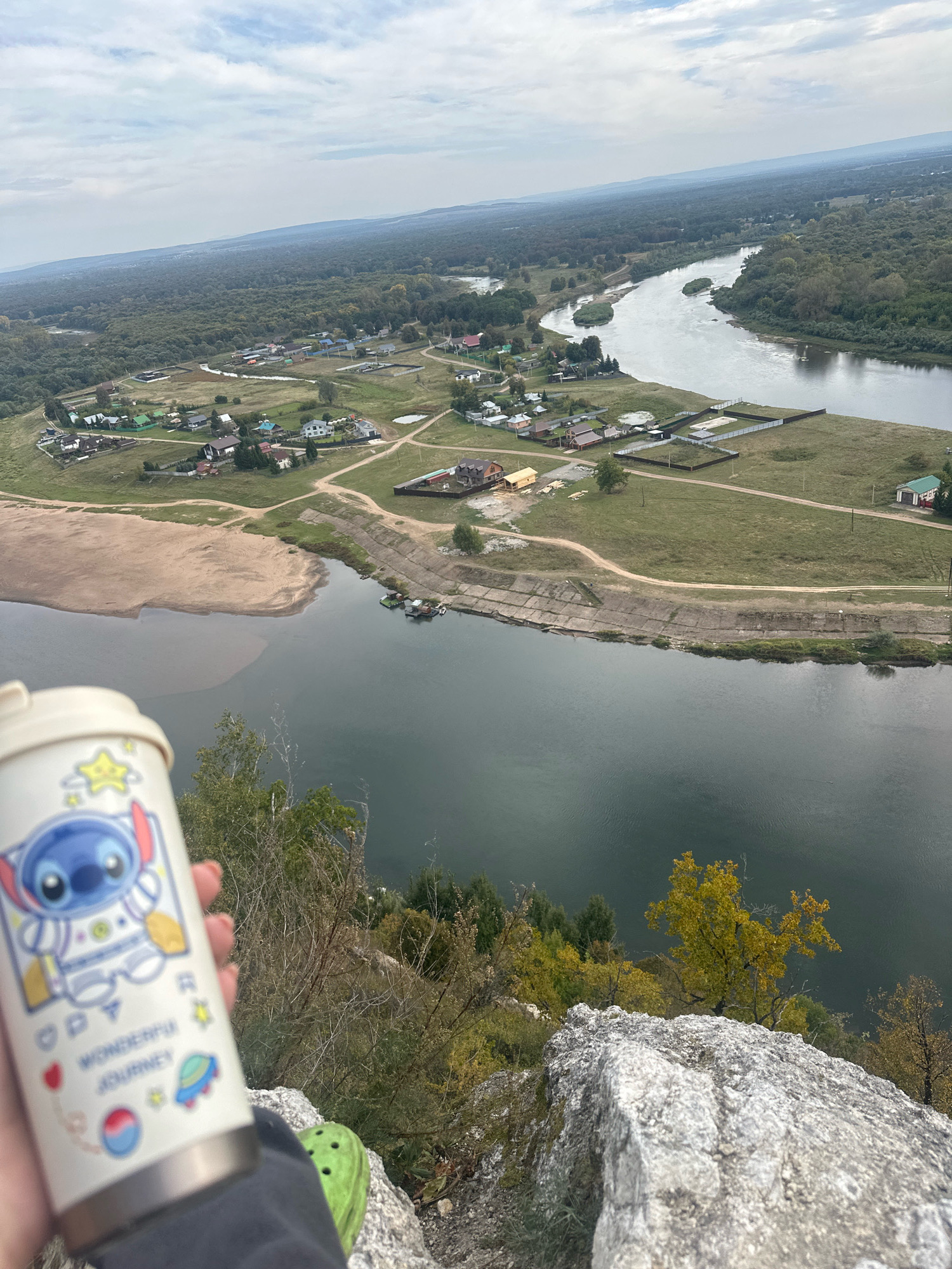 The image is taken from a high vantage point, looking down on a river and a settlement. In the foreground, a person's hand holds a white thermal mug with a cartoon character and text that says "WONDERFUL JOURNEY". Part of a green shoe is visible at the bottom right of the frame. The mug is in focus, while the background is slightly blurred.
The background shows a wide river curving through a landscape dotted with houses and trees. The settlement appears to be along the riverbank, with some areas of sandy beach. Further into the distance, the landscape is covered by a dense forest, and the sky is overcast with clouds. The terrain is hilly. This location is identified as Simskii, Russia.