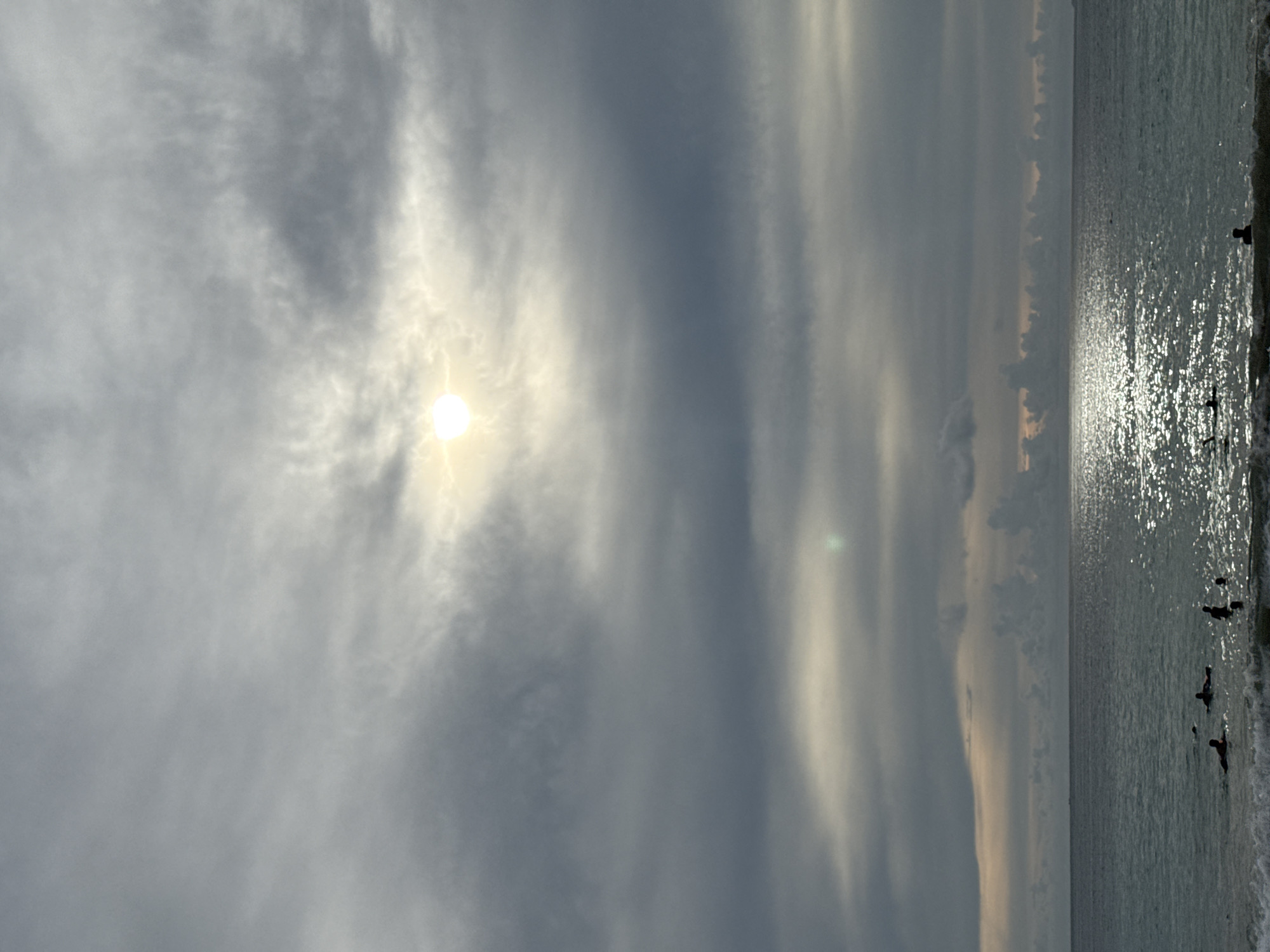 The image captures a serene ocean scene with a dramatic sky. The sun, partially obscured by wispy clouds, casts a bright, diffused light. The clouds are varied, with some appearing thin and streaky, while others are denser and darker, suggesting a dynamic atmosphere. The ocean is calm, with a gentle swell and a shimmering surface reflecting the sunlight.
In the water, several figures can be seen, appearing to be people engaging in water activities, possibly surfing or swimming. Their dark silhouettes contrast with the bright reflection on the water. The shoreline is visible on the right, with a hint of waves breaking.
The overall mood is peaceful and contemplative, with the interplay of light and shadow creating a visually striking image. The time of day appears to be either early morning or late afternoon, given the quality of the sunlight and the subdued lighting. The location, Meunasahbale, Indonesia, suggests a tropical or coastal setting. There is no visible text in the image.