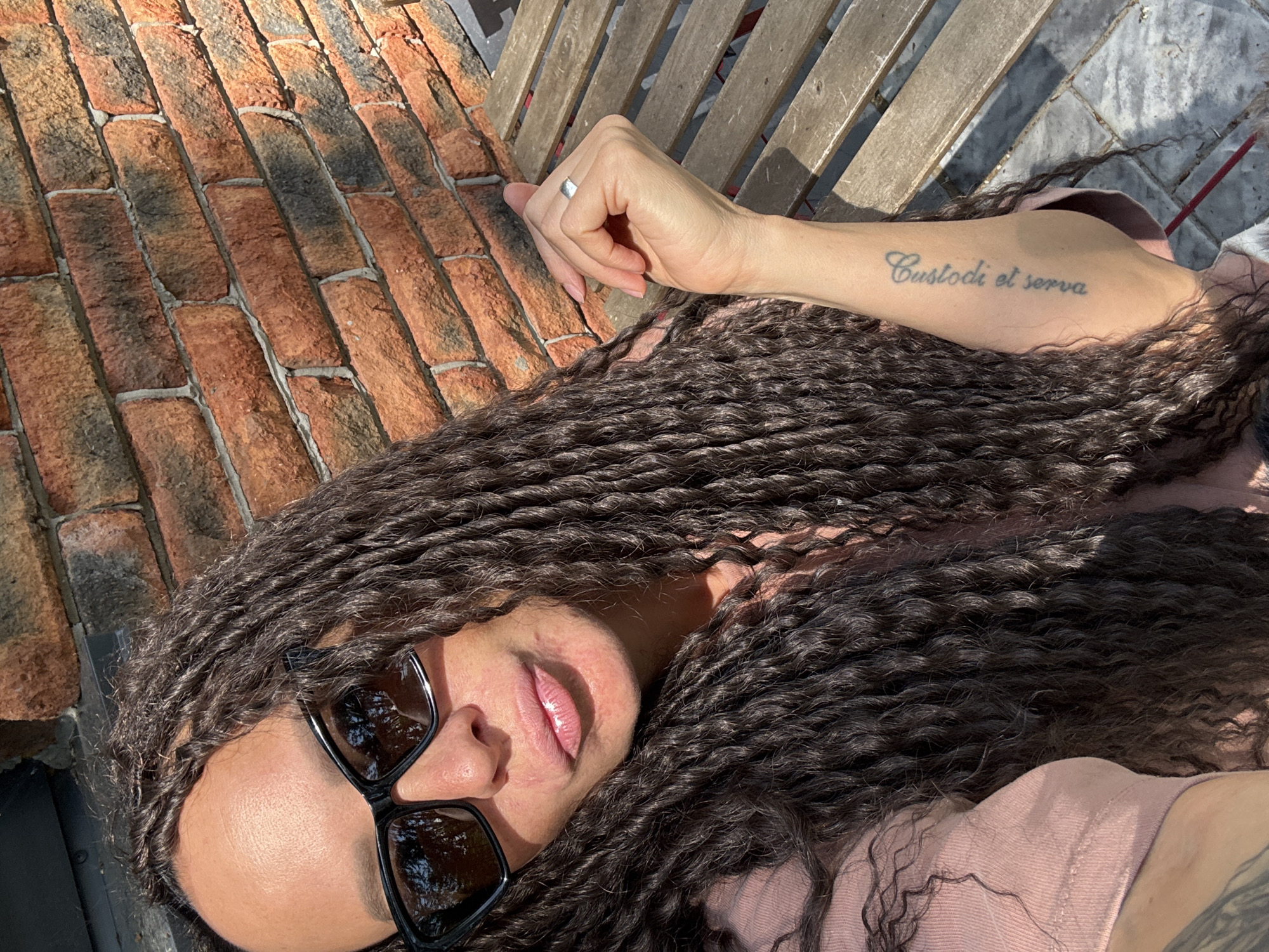 A medium-shot photo shows a person wearing sunglasses lying with their head near a brick surface and their arm extended toward some wooden slats. A silver ring is visible on the person's ring finger. A tattoo reading "Custodi et serva" is visible on the person's arm. The location is likely outdoors, judging by the sunlight and shadow. There is no information in the image that specifies that it is in Chaikovsky, Russia.