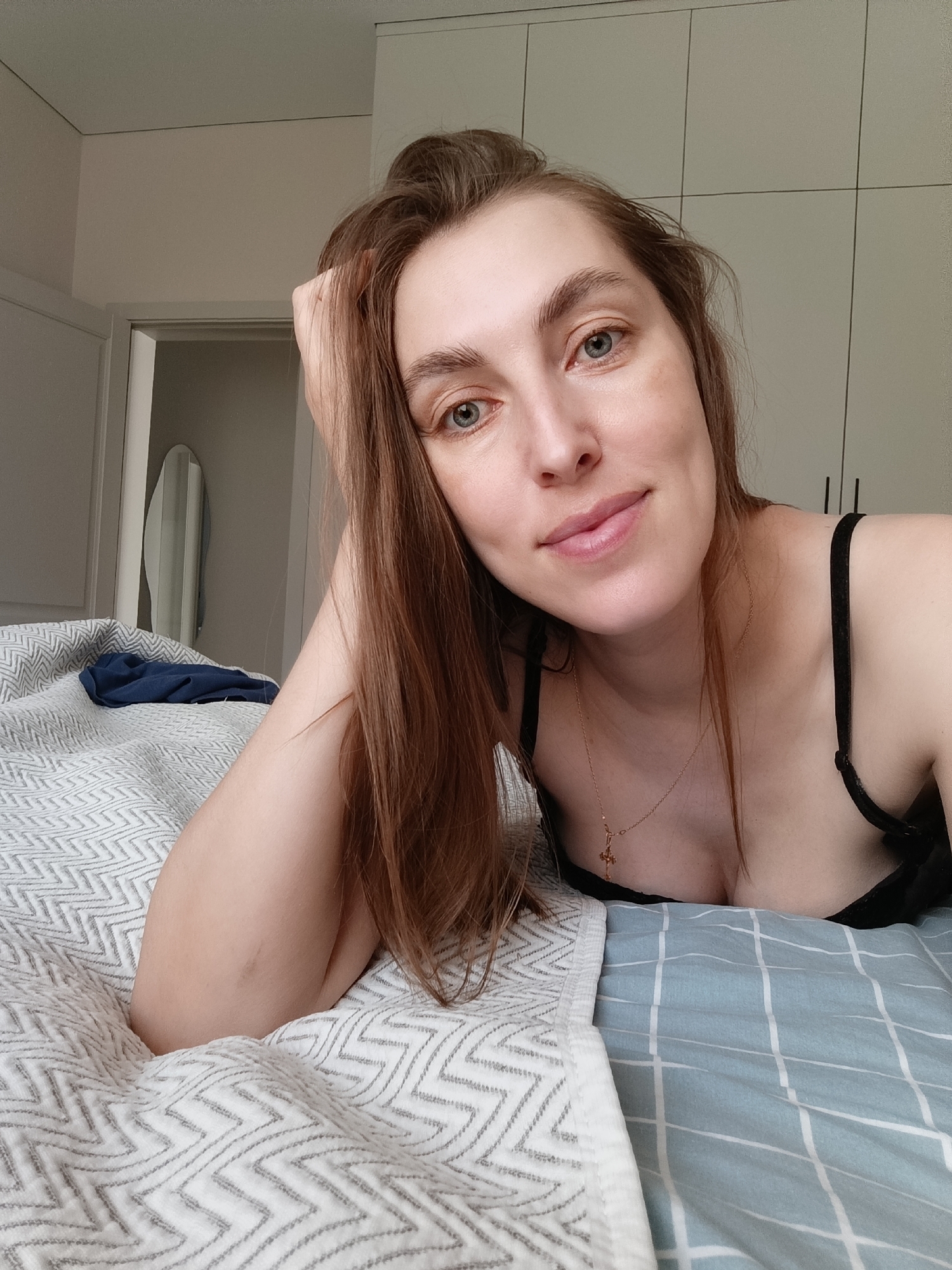 A woman with long brown hair is lying on a bed. She is wearing a black bra and a gold necklace with a cross pendant. Her head rests on her left hand. The bed has a white patterned comforter and a blue checkered sheet. In the background, there is a white cabinet and a doorway with a mirror. The image was likely taken indoors in a private residence. There is no information provided in the image that indicates the city of Chaikovsky, Russia.