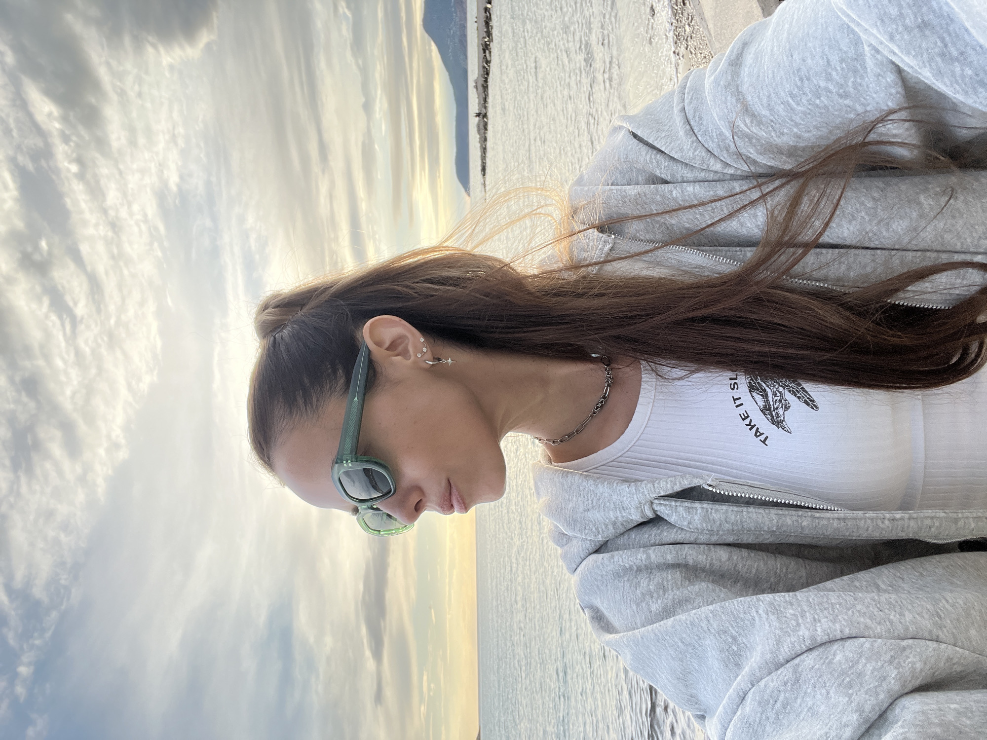 A woman with light brown hair is standing on the shore near Bar, Montenegro, facing the sea. She is wearing sunglasses, a white top, a silver chain necklace, and a gray zip-up hoodie. The sky has scattered clouds and a golden hue from the setting sun. In the background is the coastline of Montenegro and a calm sea.