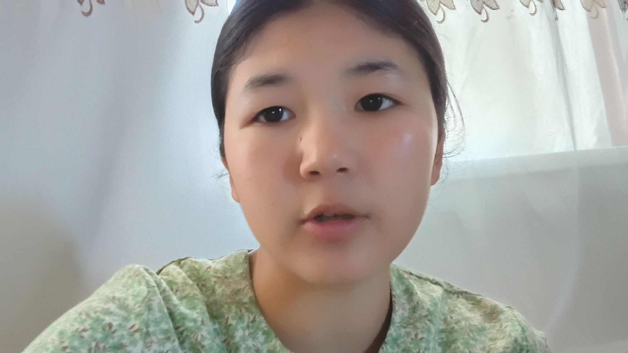 A close-up, head-and-shoulders portrait shows a young East Asian female, positioned centrally, looking directly at the camera. She has dark hair pulled back, revealing her forehead and ears. Her eyes are dark and open, and her lips are slightly parted. She is wearing a light green collared shirt with a subtle floral pattern. The background consists of a light-colored wall or curtain, with a decorative element near the top that appears to be a fabric trim with an embroidered pattern. The lighting is soft and diffused, with a slight sheen on her face, suggesting it might be natural light. The image appears to be taken indoors, possibly in a residential setting. There is no information to suggest the location is in Uchkun, Kyrgyzstan.